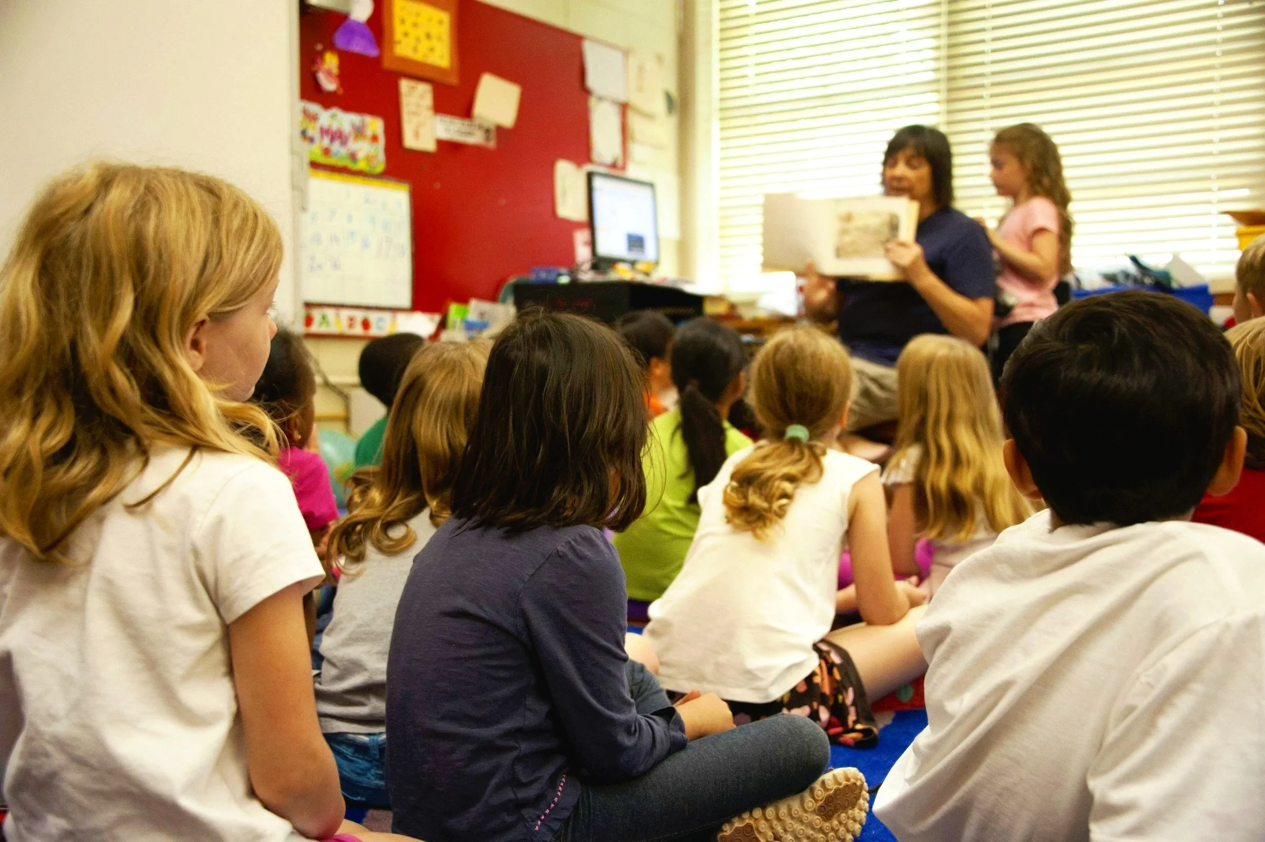 a group of children at school listening to a teacher read a book to the class
