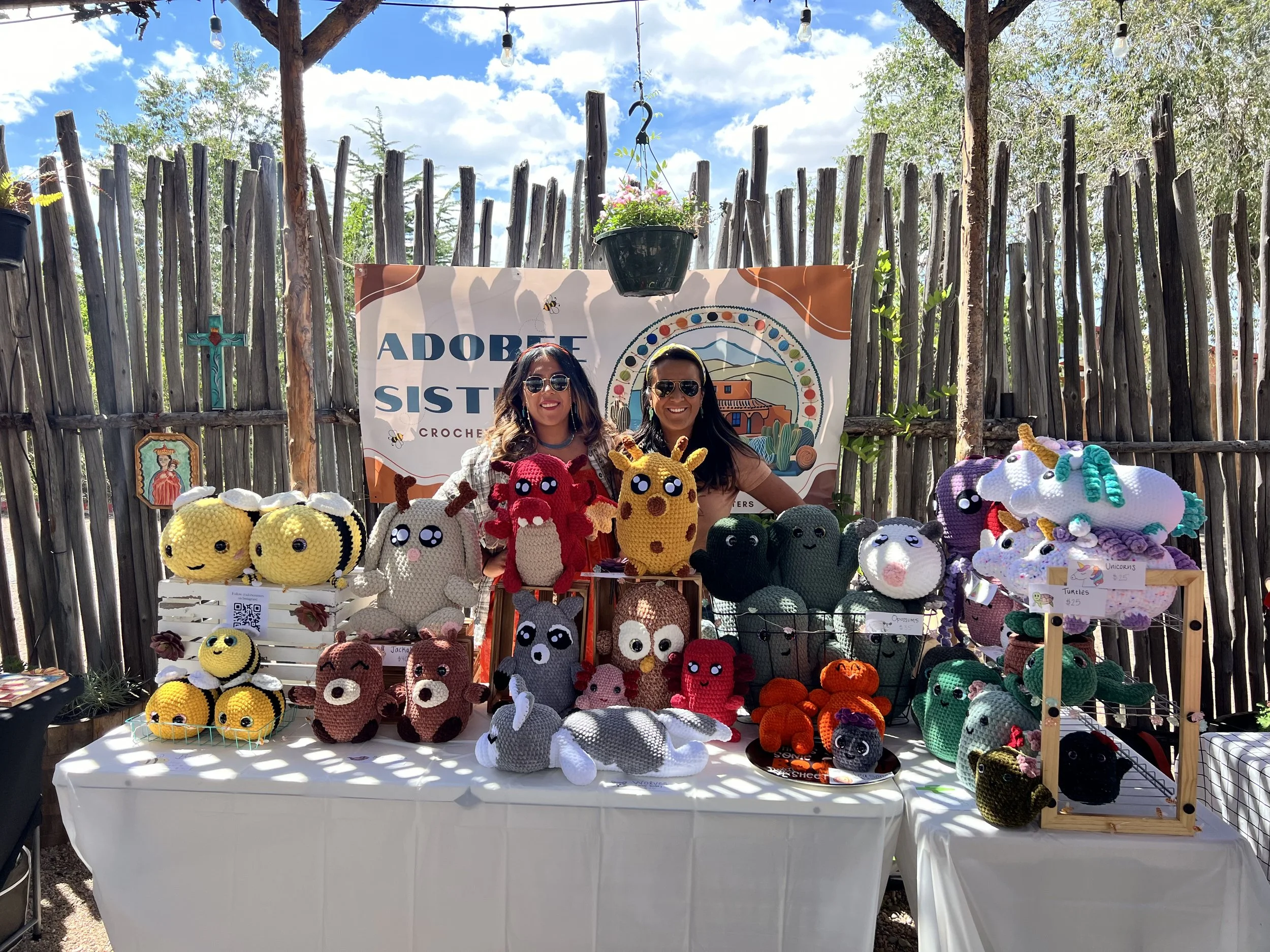 the two adobee sisters standing and smiling in front of their booth with a collection of crochet animals.