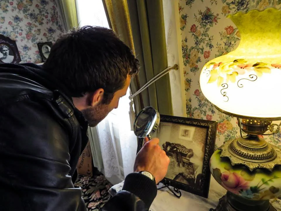 A person examining a framed photograph with a magnifying glass next to a vintage lamp on a floral wallpaper background.