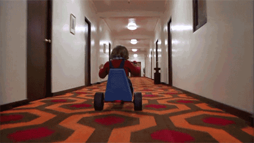 Child riding a tricycle down a hotel hallway with patterned carpet.