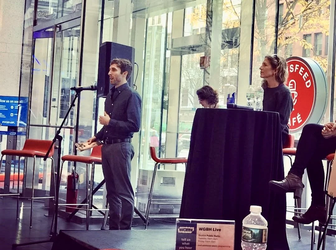 A man speaking into a microphone at a public event inside a cafe or restaurant, with two women seated behind him, one at a table and another on a high chair, near large glass windows showing a city street outside.