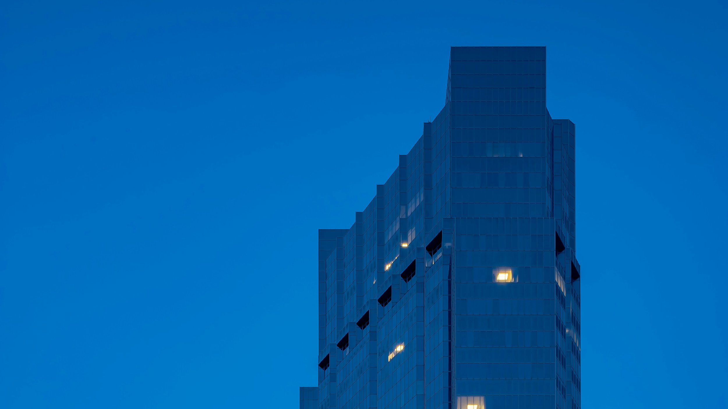 A modern high-rise building with a glass facade is silhouetted against a deep blue sky during twilight. A few windows are illuminated with warm yellow light, creating a contrast with the cool tones of the building and sky. London.