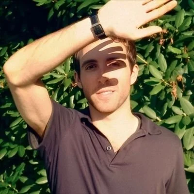 Young man with short brown hair, wearing a dark gray polo shirt and a black wristwatch, standing outdoors in front of green leafy bushes, shielding his eyes from the sun with his right hand.
