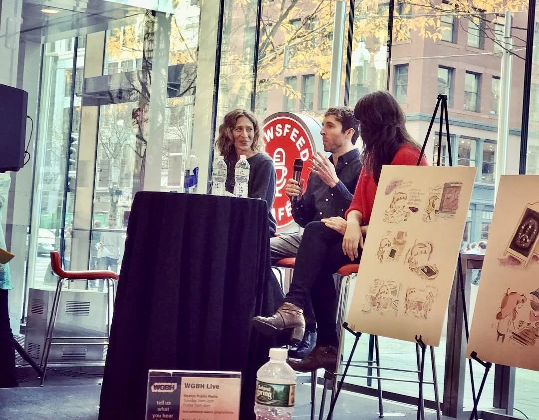 Three people are seated at a table during a panel discussion or interview inside a glass-walled building with trees visible outside. A woman with wavy hair and a black jacket, a young man with short hair holding a microphone, and another woman with long dark hair and a red top are engaged in conversation. Several posters or artwork are displayed on easels nearby, and a water bottle and some signs are on the table and floor.