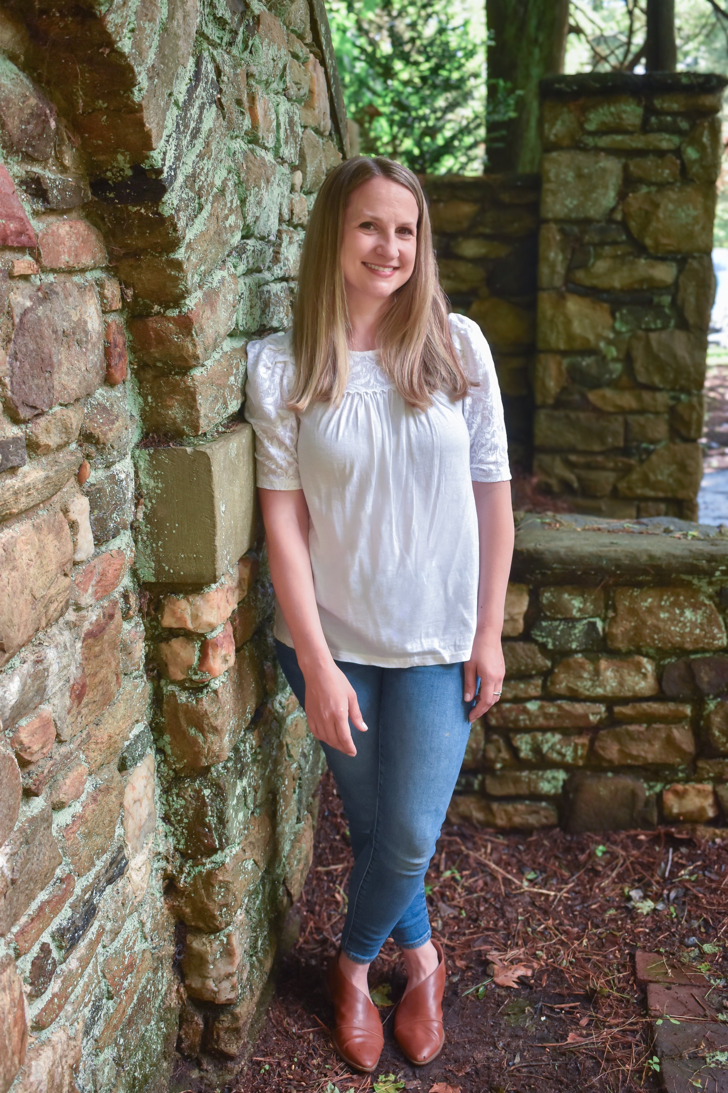 A woman with long blonde hair smiling, standing outdoors near a stone wall, wearing a white blouse, blue jeans, and brown shoes.