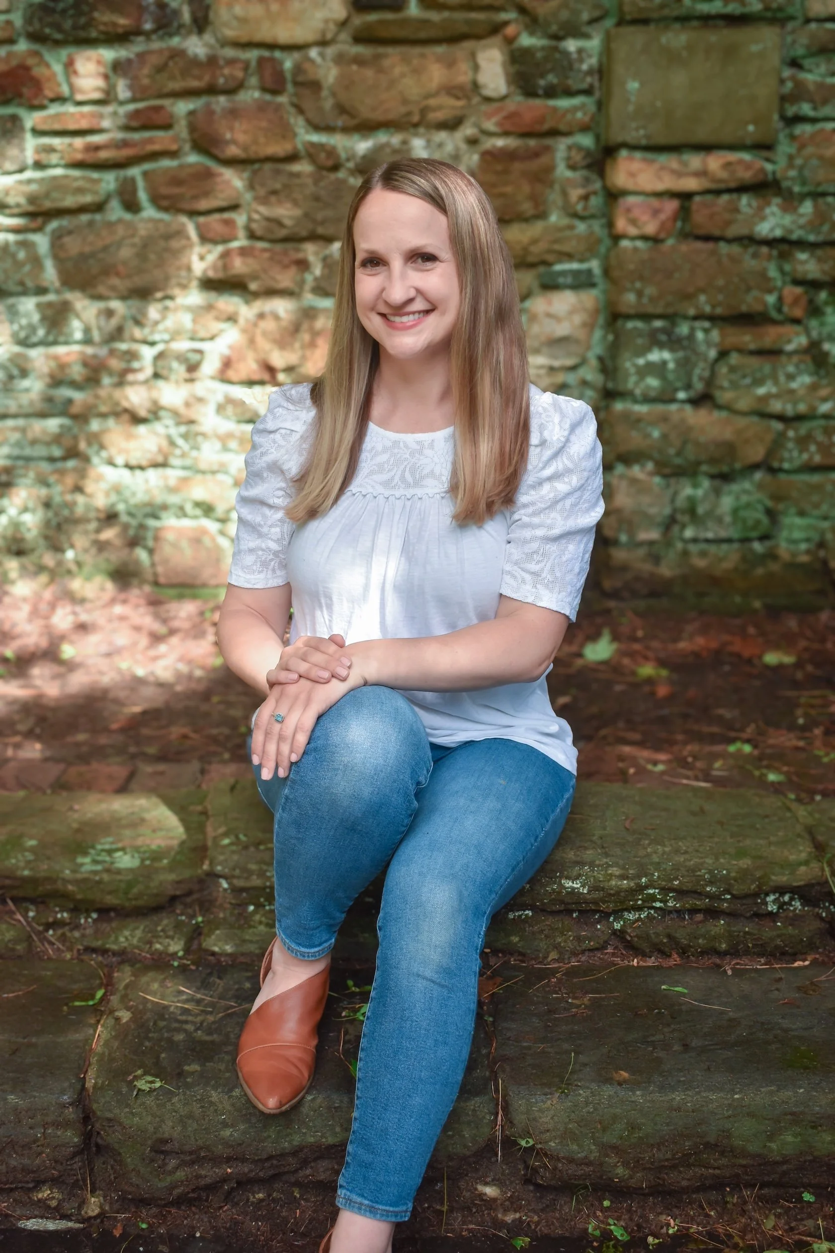 A woman sitting on mossy stone steps outdoors, in front of a stone wall. She has long light brown hair, is wearing a white blouse with short puffed sleeves, blue jeans, and tan shoes, and is smiling at the camera.