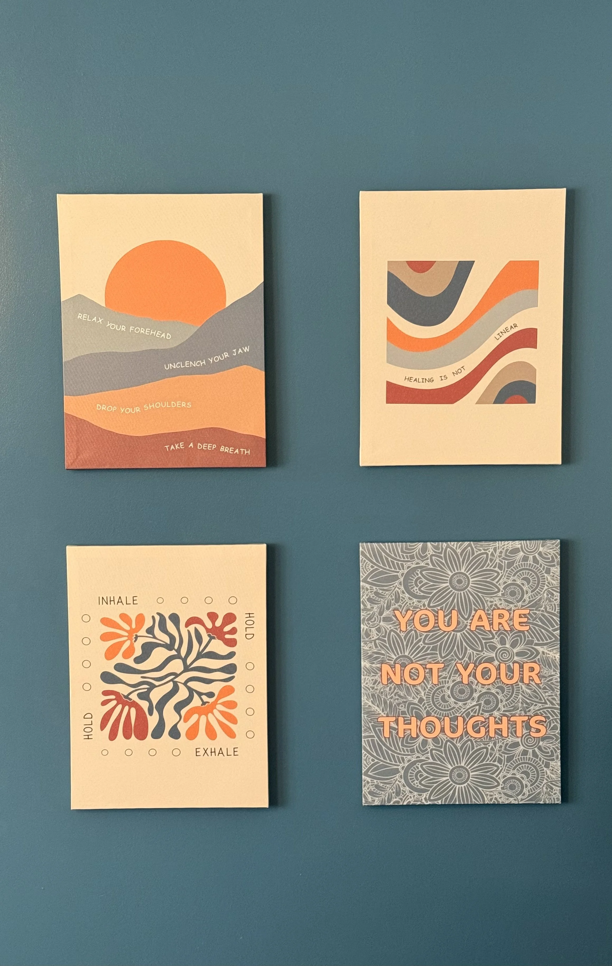 Four motivational and wellness-themed posters on a teal wall. The top left poster features a stylized sunrise and mountain landscape with the words, "Relax your forehead, Unclench your jaw, Drop your shoulders, Take a deep breath." The top right poster has abstract, wavy shapes with the phrase, "Healing is not linear." The bottom left poster depicts a simple, abstract tree with the words, "Inhale, Hold, Exhale, Hold." The bottom right poster displays the phrase, "You are not your thoughts," over a floral background.