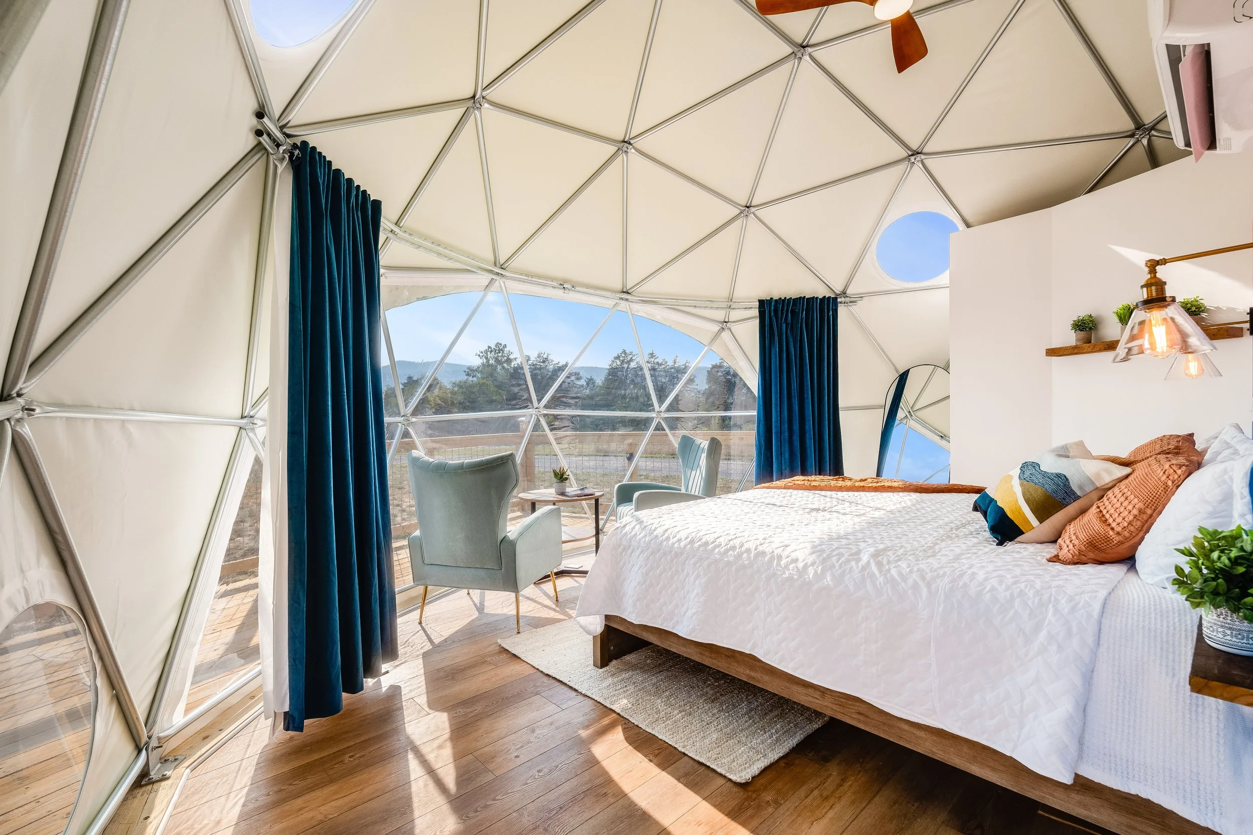 Interior of a geodesic dome with a bed, two chairs, and a view outside through triangular windows. The room has wooden flooring, modern decor, and blue curtains.