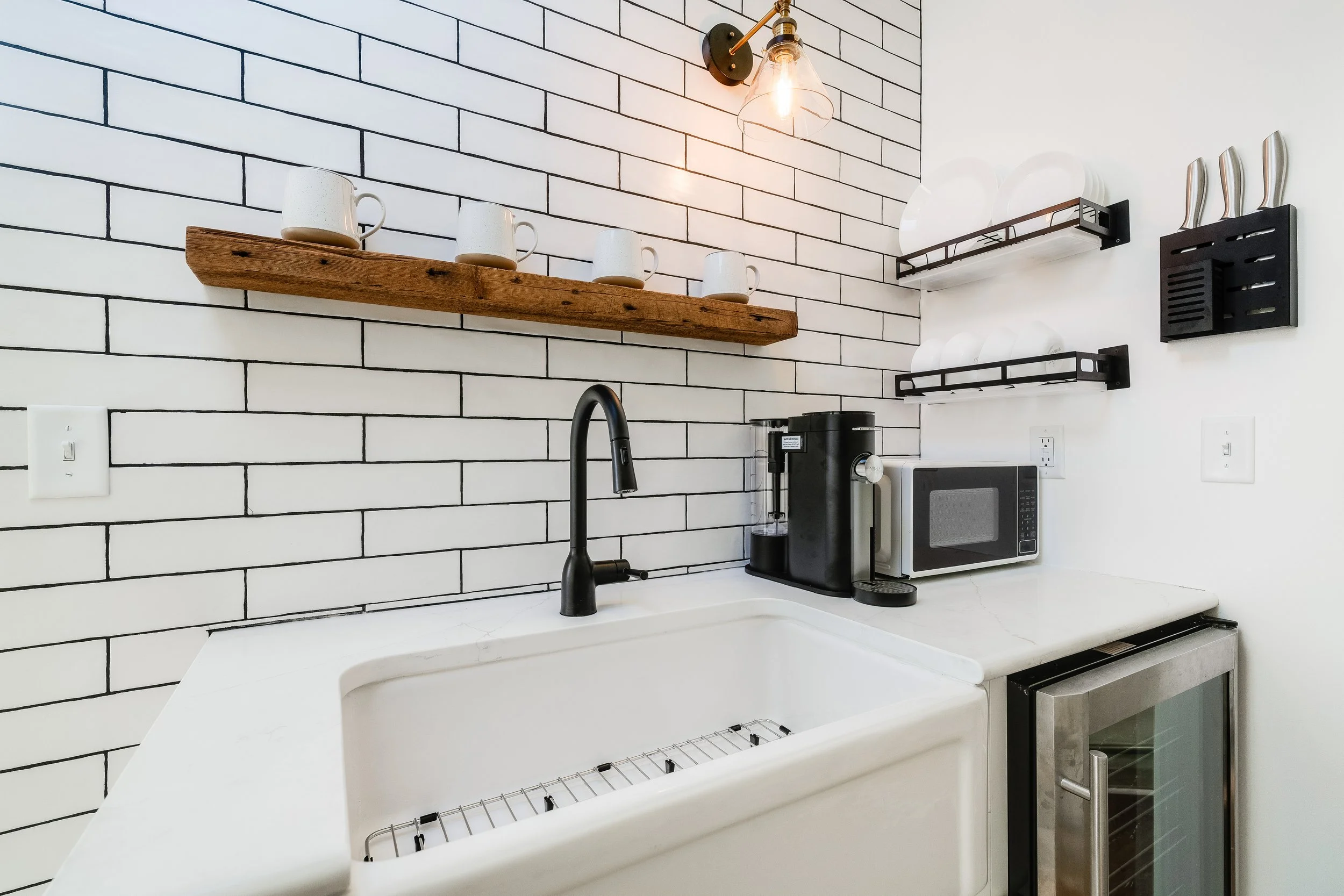 Modern kitchen with a white subway tile backsplash, farmhouse sink, black faucet, wooden shelf with white mugs, wall-mounted racks with plates, countertop microwave, coffee maker, and a built-in mini-fridge below.
