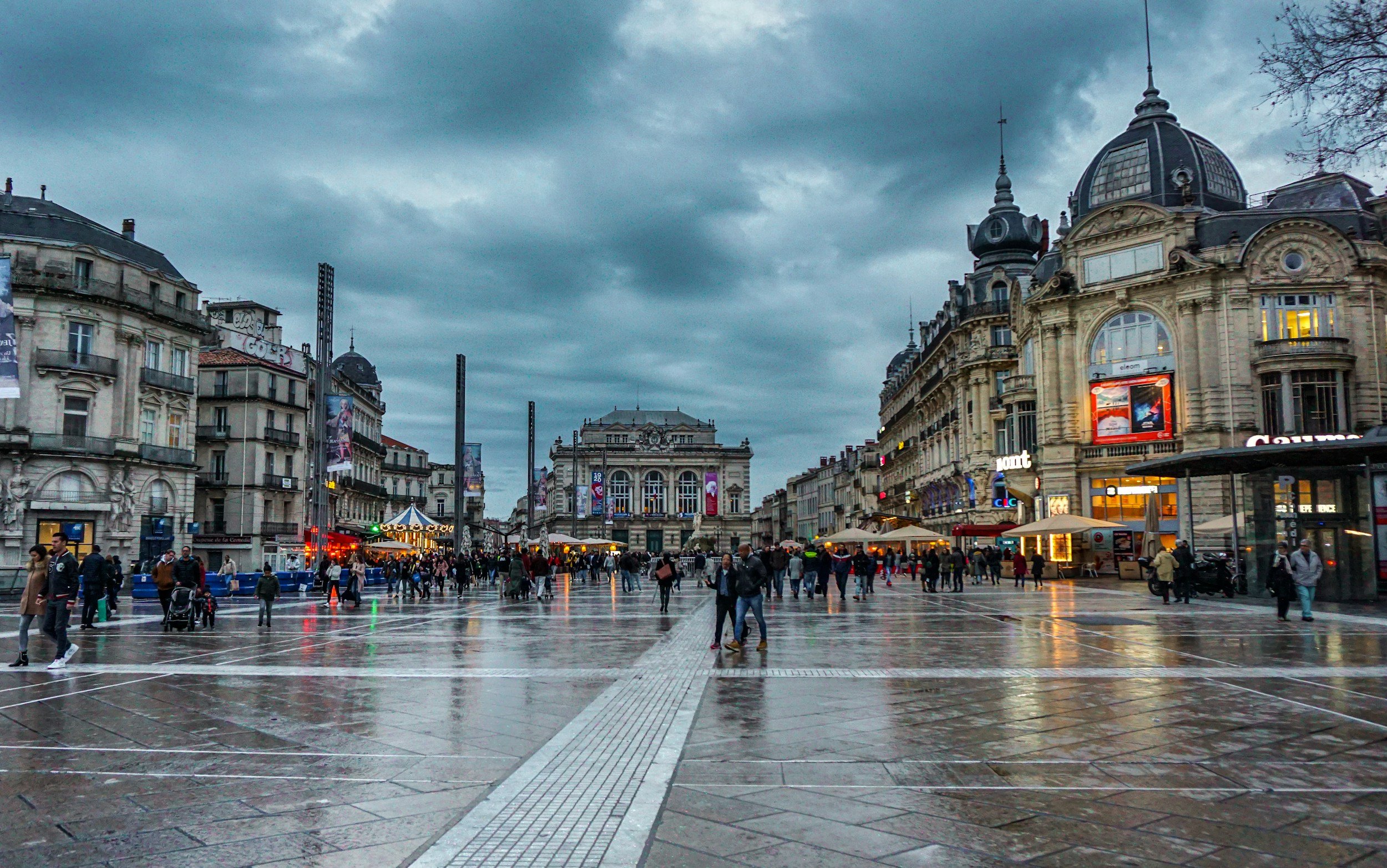 Place de la Comédie à Montpellier avec des piétons, bâtiments historiques et ciel nuageux.