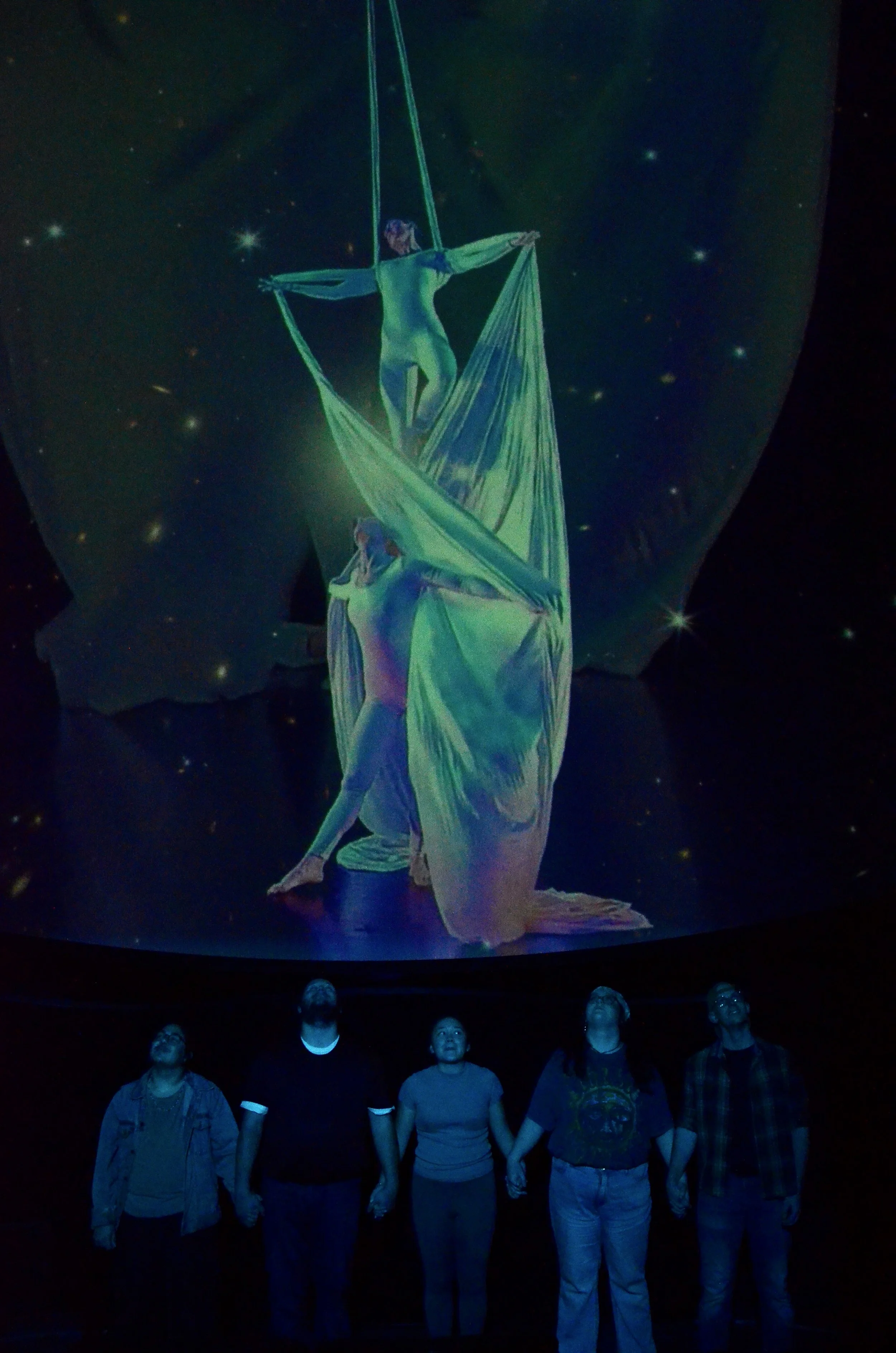 Five people holding hands and looking upwards at a large digital projection that depicts two human figures performing acrobatic dance inside a starry sky background.