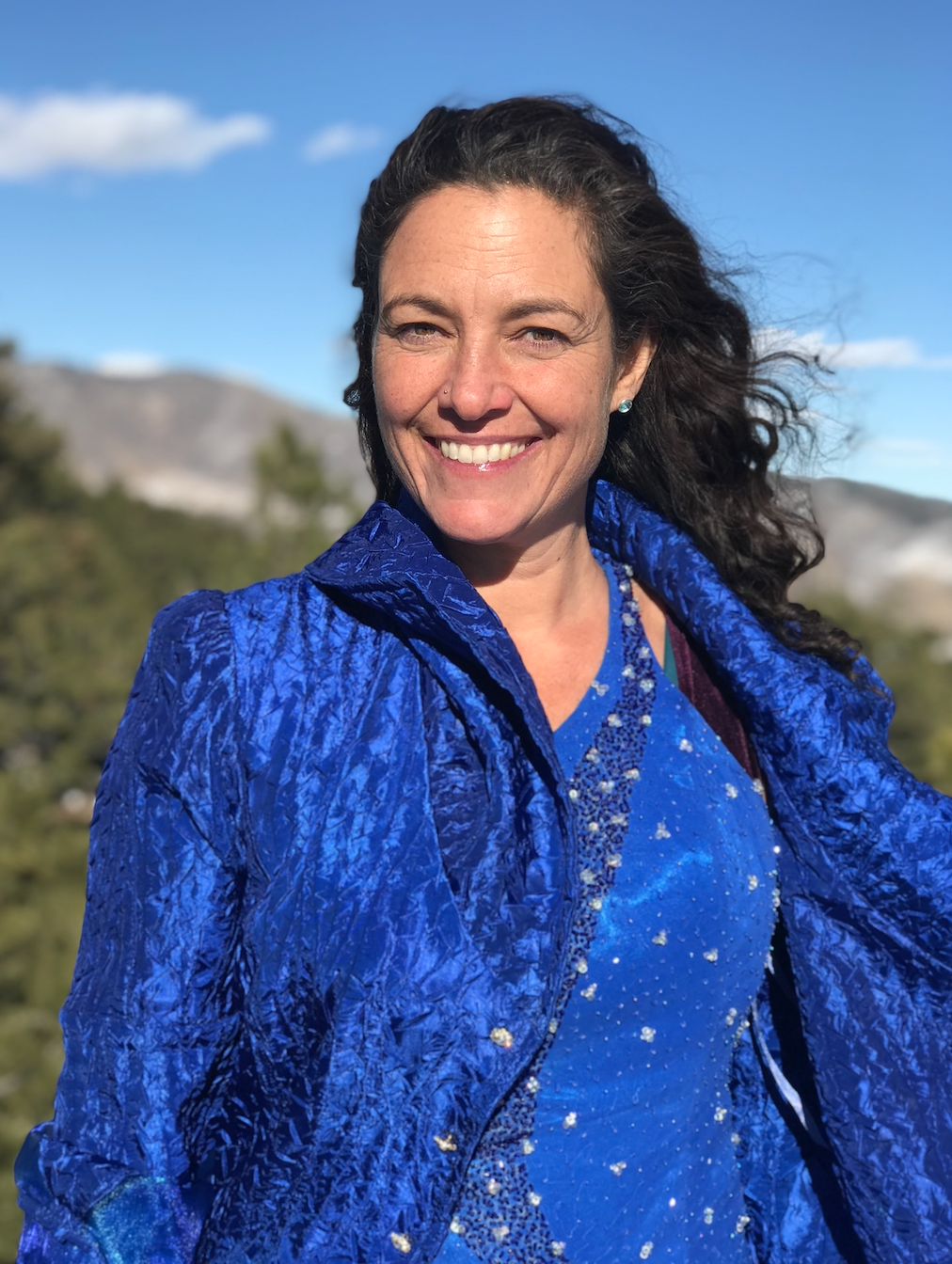 Smiling woman with dark wavy hair wearing a bright blue jacket outdoors under clear blue sky with hills and trees in the background.