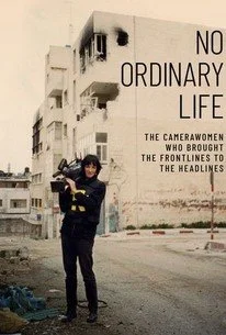 A person standing with a camera on a city street with a building in the background. The building has signage that reads 'NO ORDINARY LIFE.'