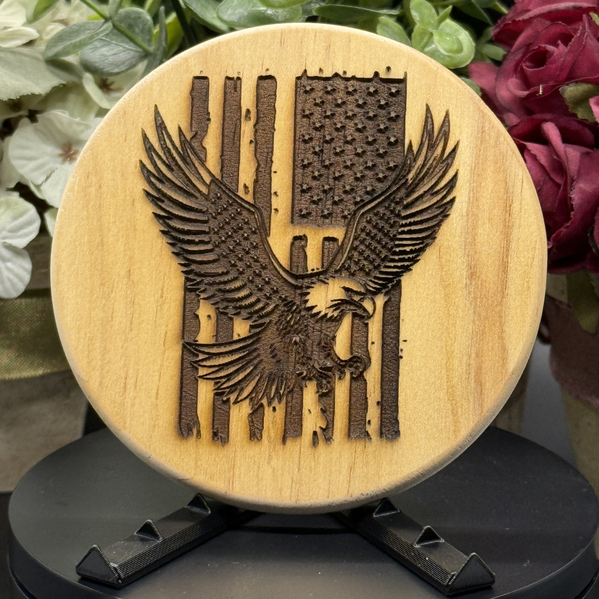 Round pine wood coaster with laser-engraved American flag and flying eagle design, displayed on a black stand with floral background