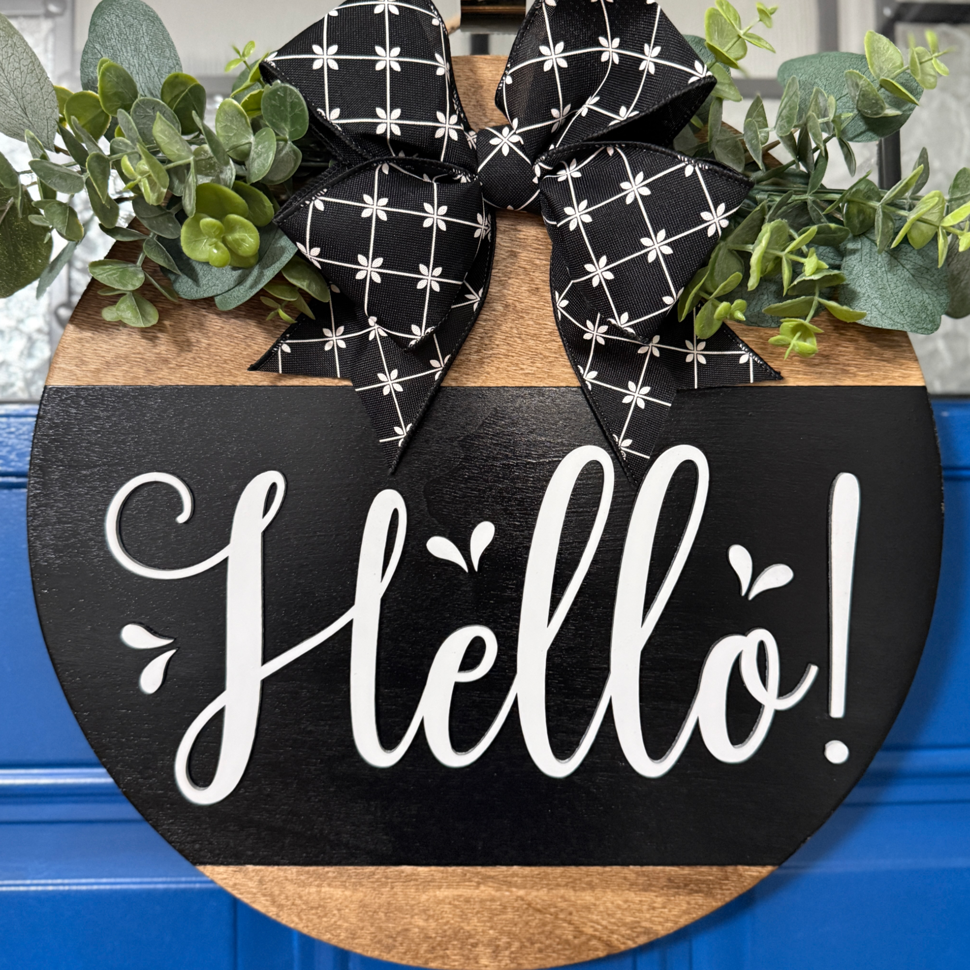 Black and natural wood round door hanger with white Hello lettering, greenery accents, and black patterned bow on blue front door