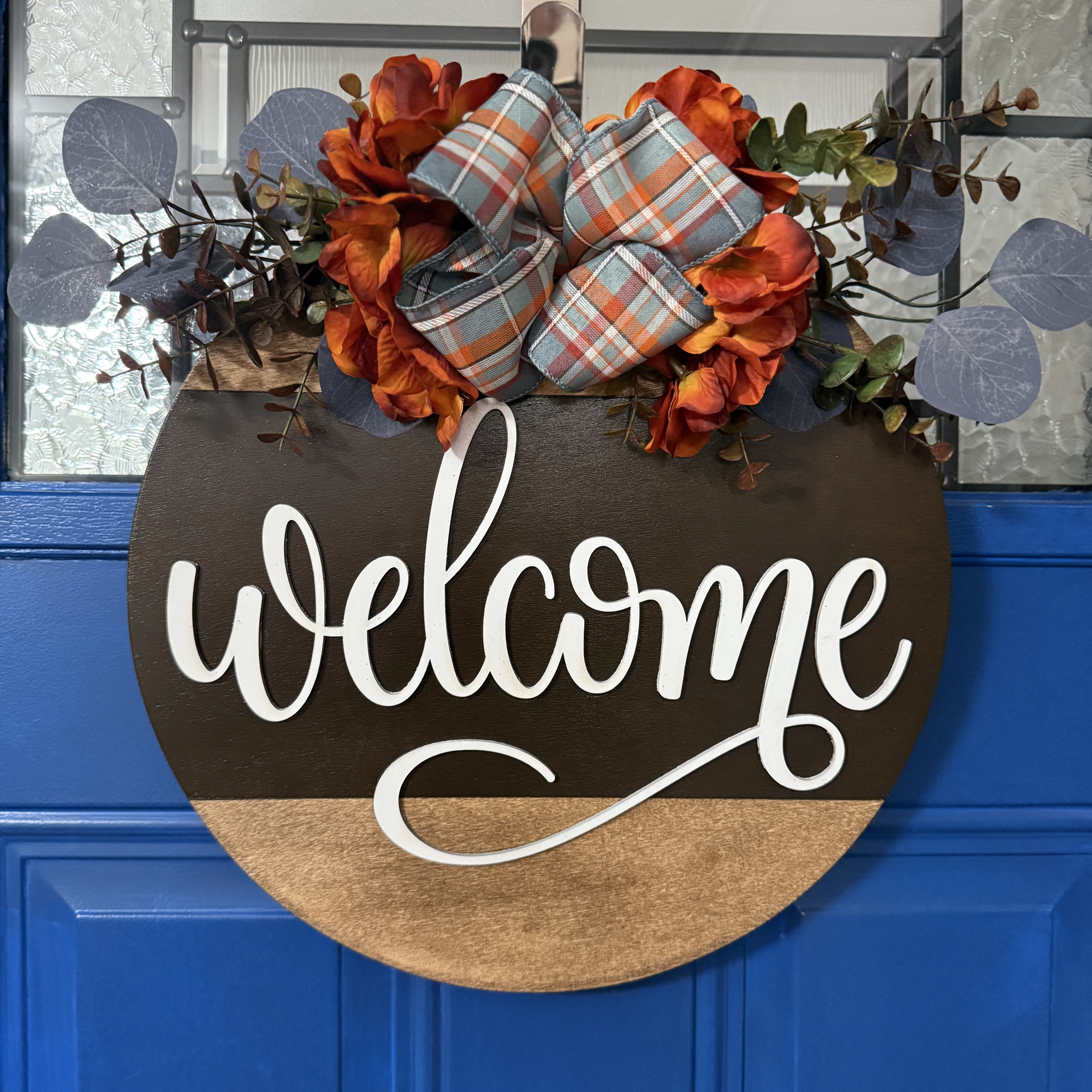 Front view of round fall welcome door hanger with plaid bow, orange florals, and greenery on rustic wood background