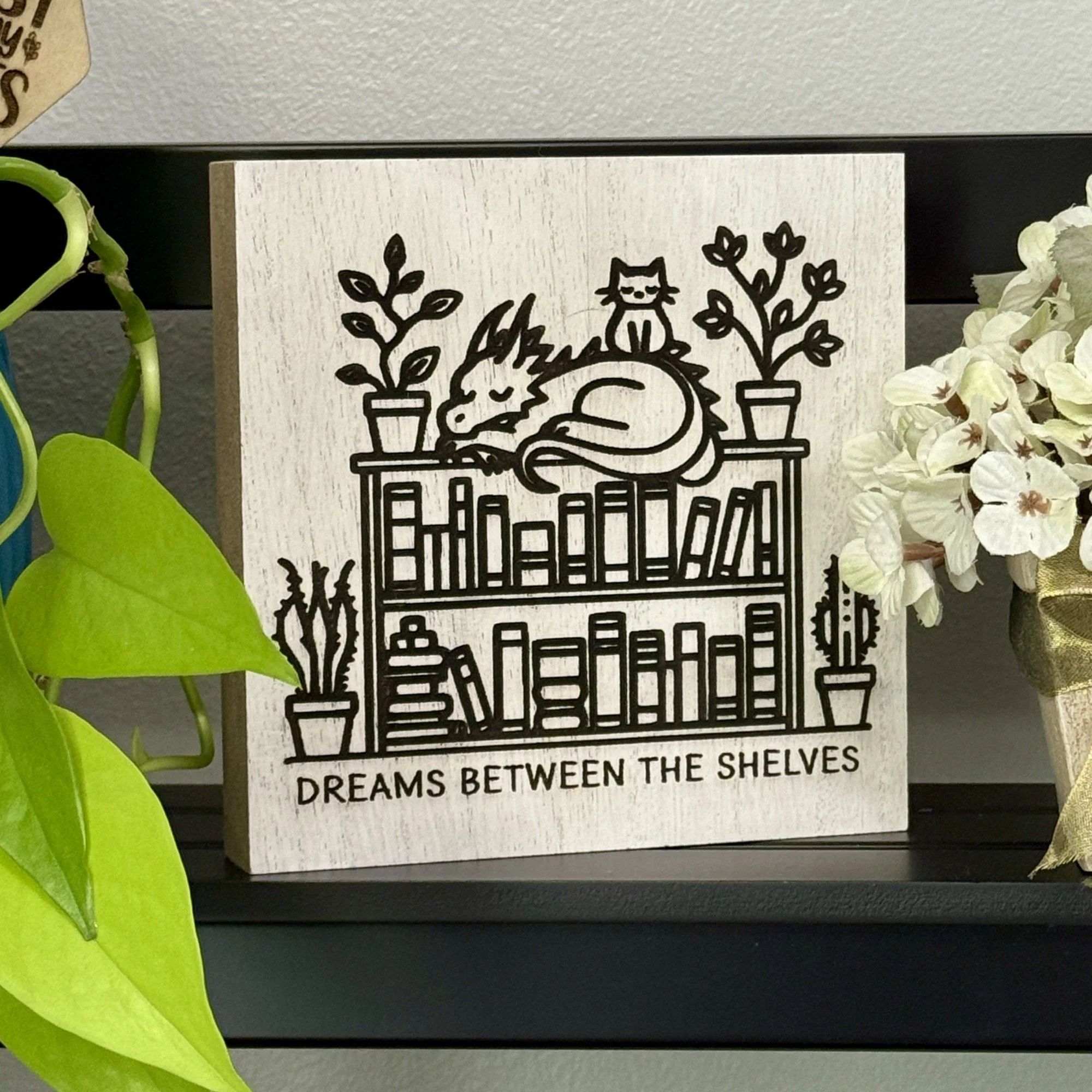 White-washed wood shelf sitter engraved with a sleeping dragon atop a bookshelf next to a cat and potted plants, displayed on a black shelf with greenery and white floral accents.