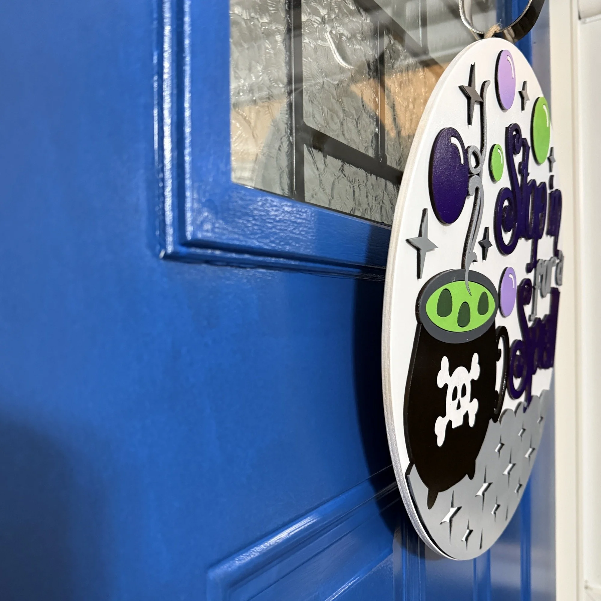 Side view of “Stop in for a Spell” Halloween door hanger with black cauldron, green brew, purple bubbles, and silver stars on a white background, hanging on a blue front door.