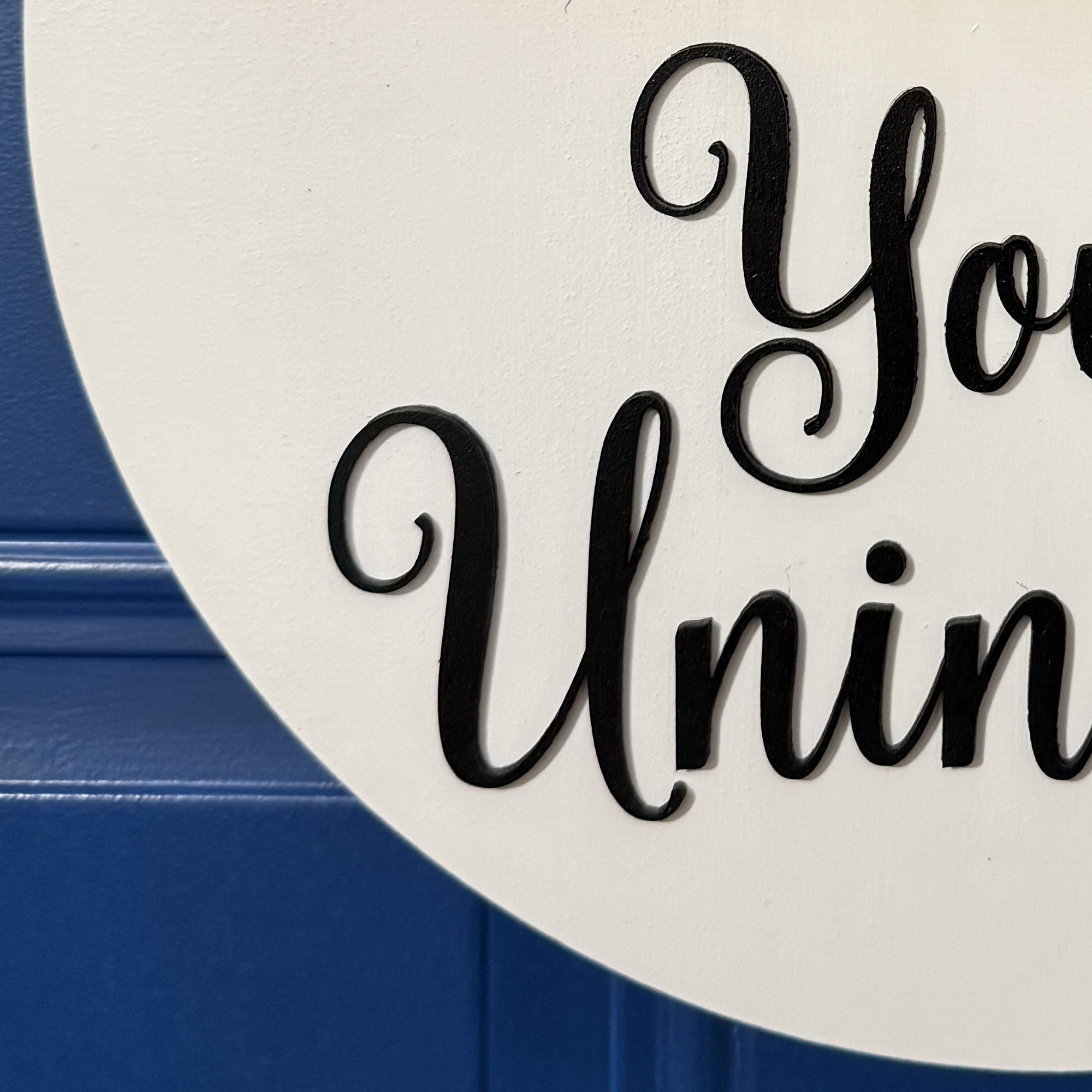 Close-up of raised black script lettering reading “You’re Uninvited” on round white wooden door hanger.