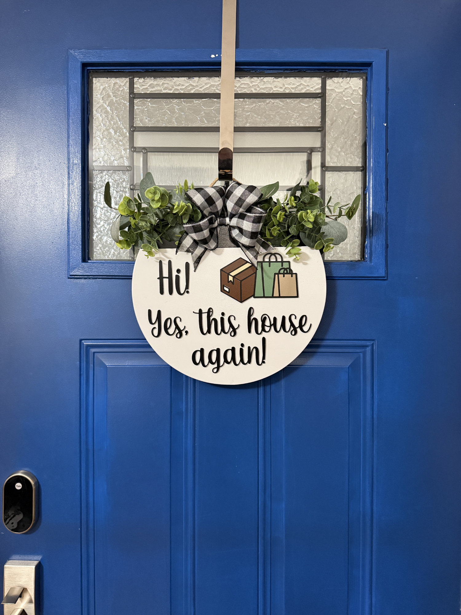 Full view of blue front door with round white “Hi! Yes, This House Again” wooden door hanger featuring dimensional lettering, greenery, and buffalo plaid bow.