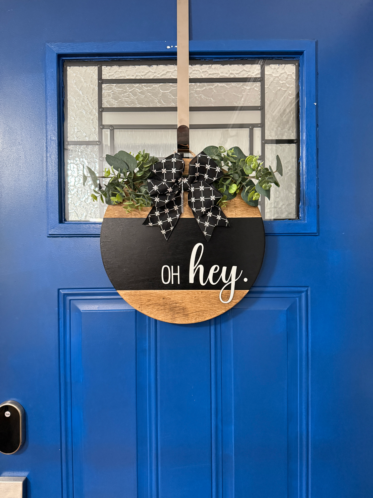 Full front door display featuring black oh hey door hanger with greenery accents and patterned ribbon bow