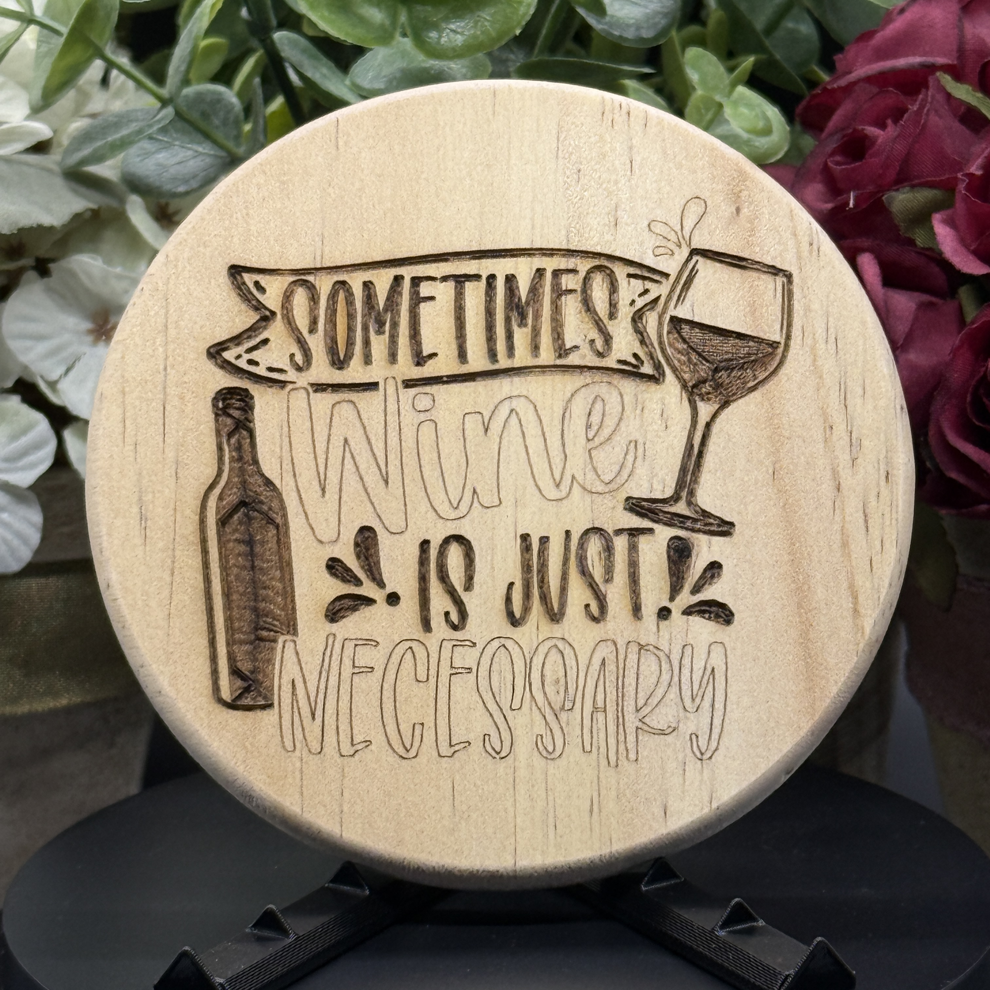 Round pine wood coaster with engraved design of a wine bottle and glass and the phrase ‘Sometimes Wine Is Just Necessary,’ shown with floral background