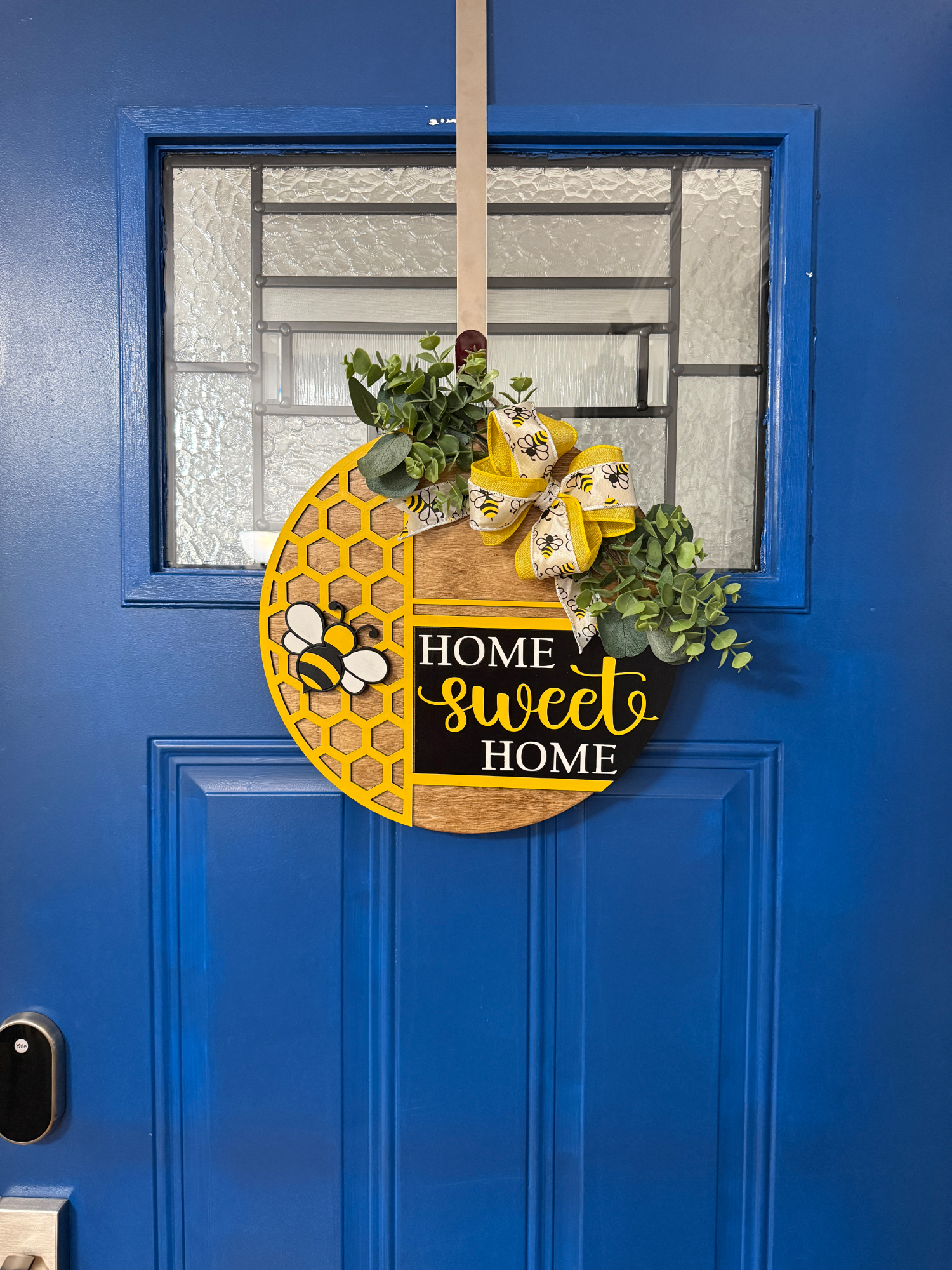 Front door display of honeycomb-themed round wood door hanger with bees, greenery, burlap bow, and “Home Sweet Home” sign on a blue door