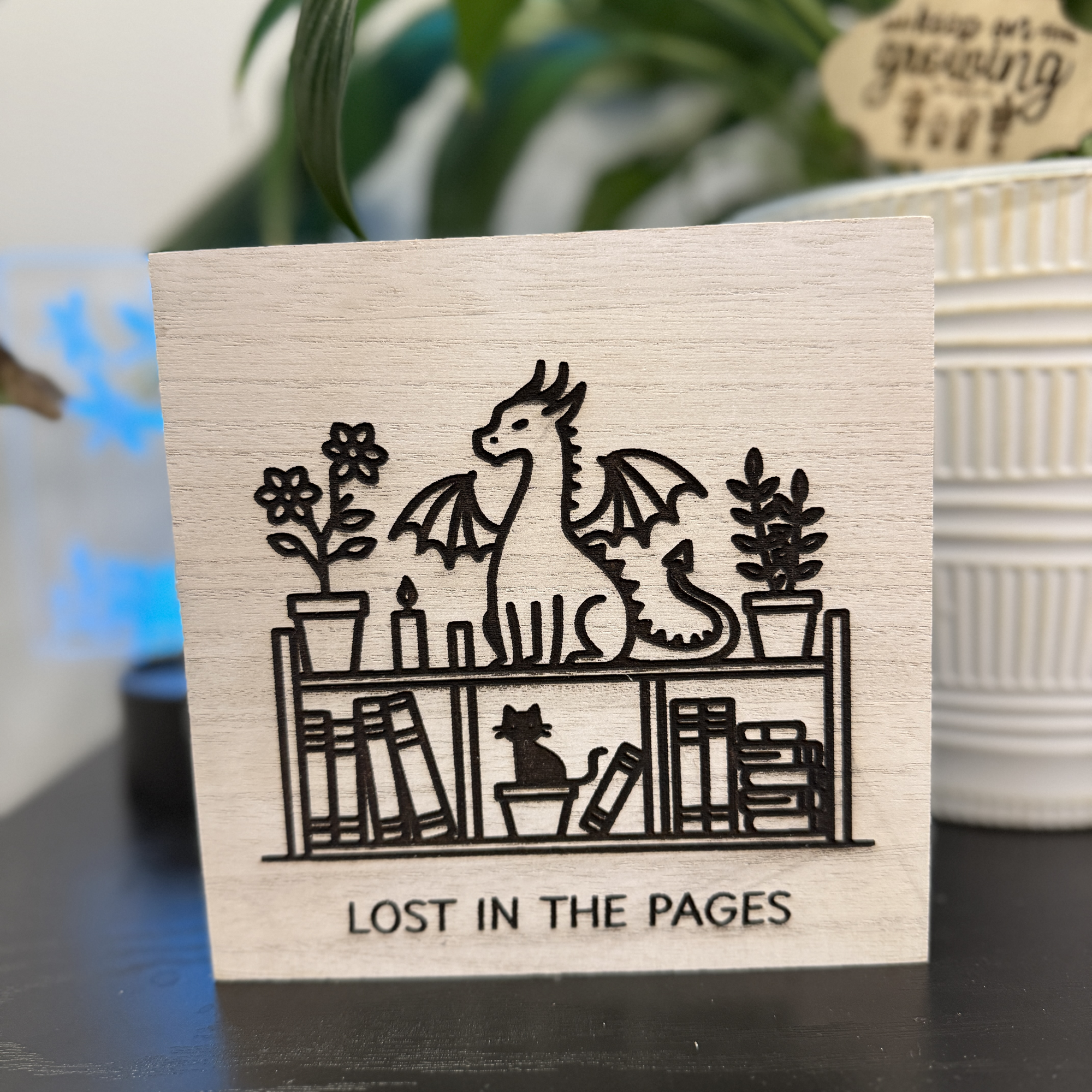 Front view of 6x6 wooden shelf sitter featuring engraved dragon and plants on a bookshelf with text reading “Lost in the Pages”