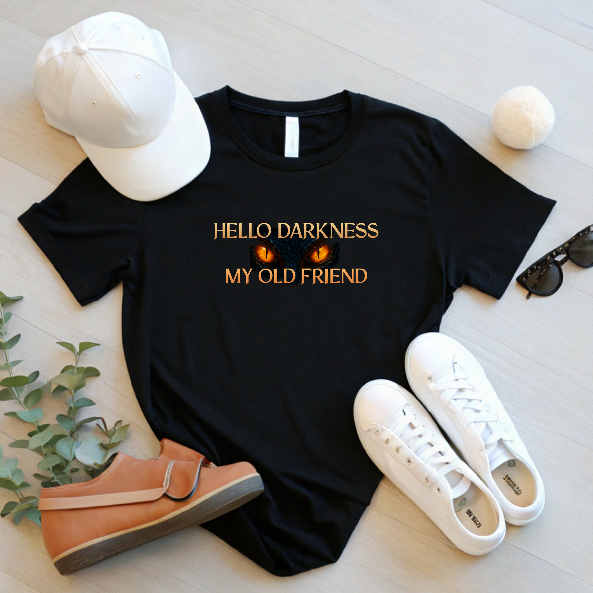 Flat lay of black “Hello Darkness My Old Friend” shirt with white hat, white shoes, sunglasses, and greenery on a light floor.
