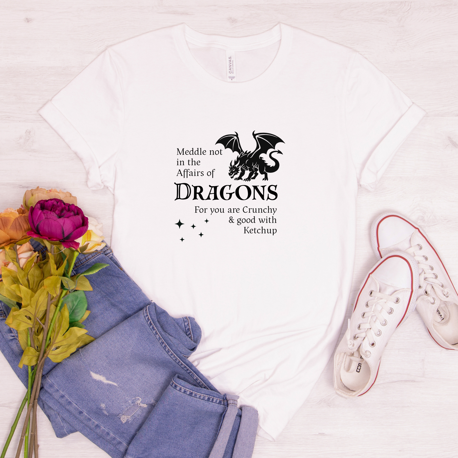 White dragon quote t-shirt with black design, displayed with flowers, jeans, and shoes on a light background.
