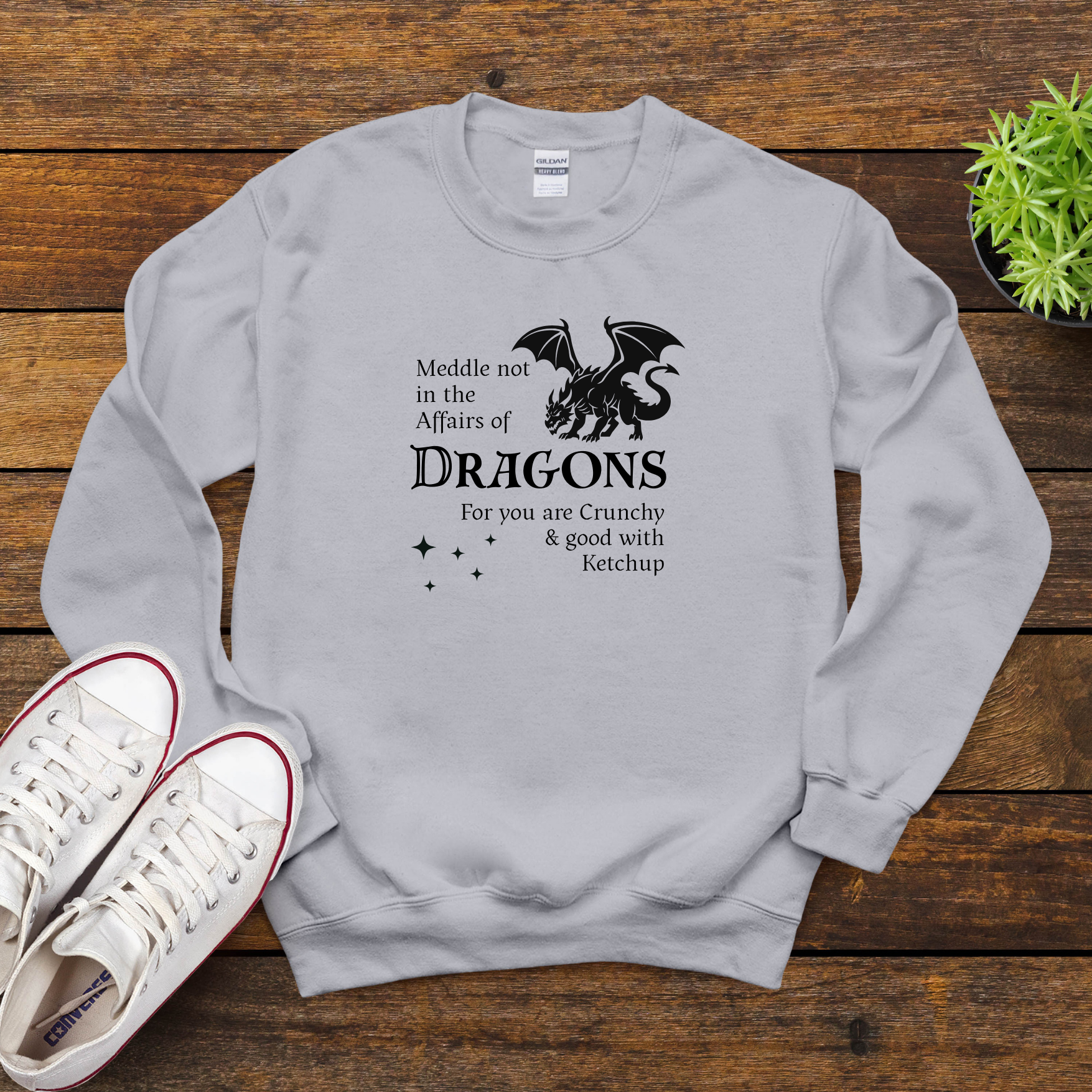 Flatlay of light gray dragon sweatshirt on wood background with shoes, featuring white dragon graphic and the phrase “Meddle not in the affairs of dragons.”
