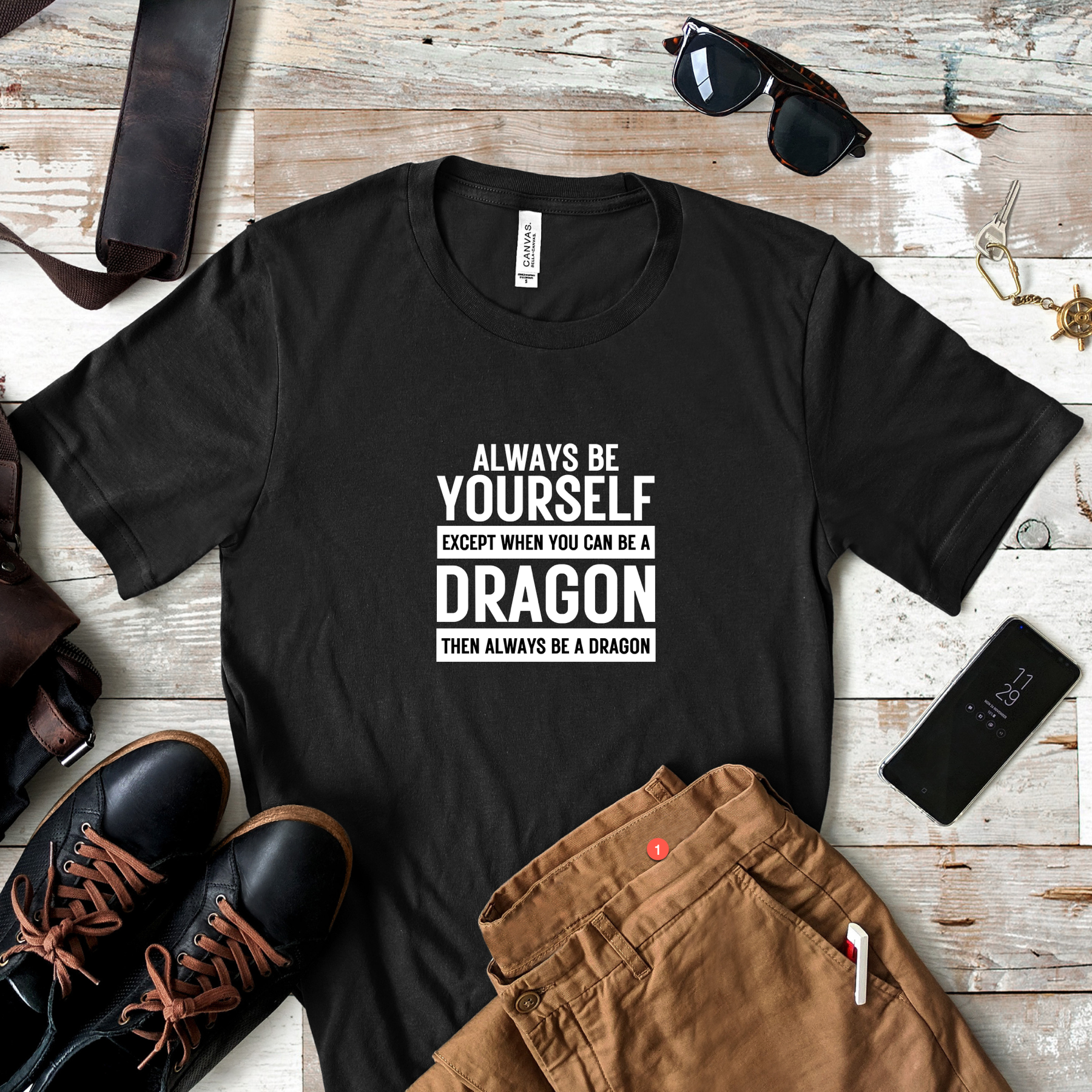 Flatlay of black unisex T-shirt with bold white text reading “Always Be Yourself Except When You Can Be a Dragon Then Always Be a Dragon,” styled with boots, jeans, and a phone.
