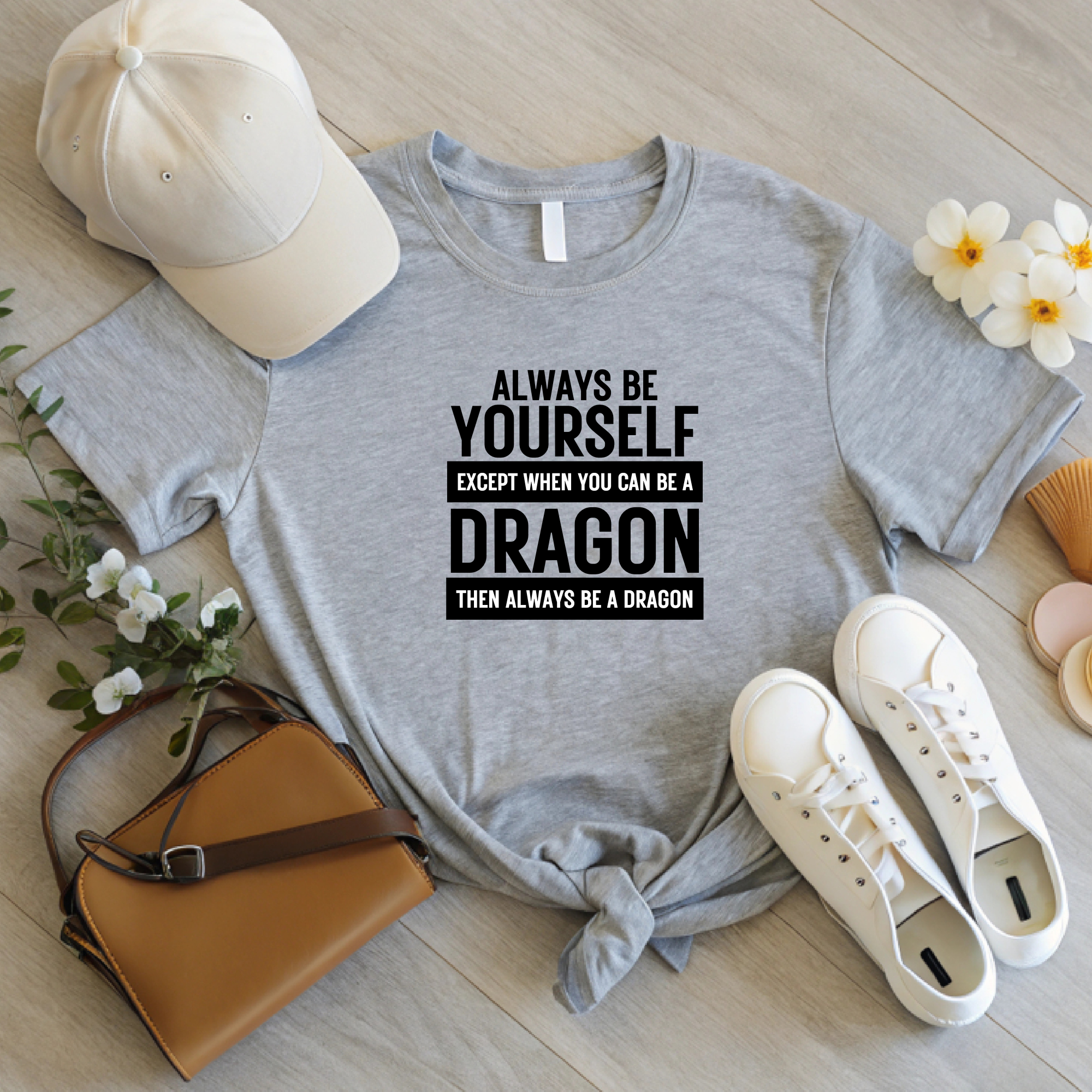 Light gray unisex T-shirt with black dragon quote design, displayed on a wood surface with hat, bag, and shoes.