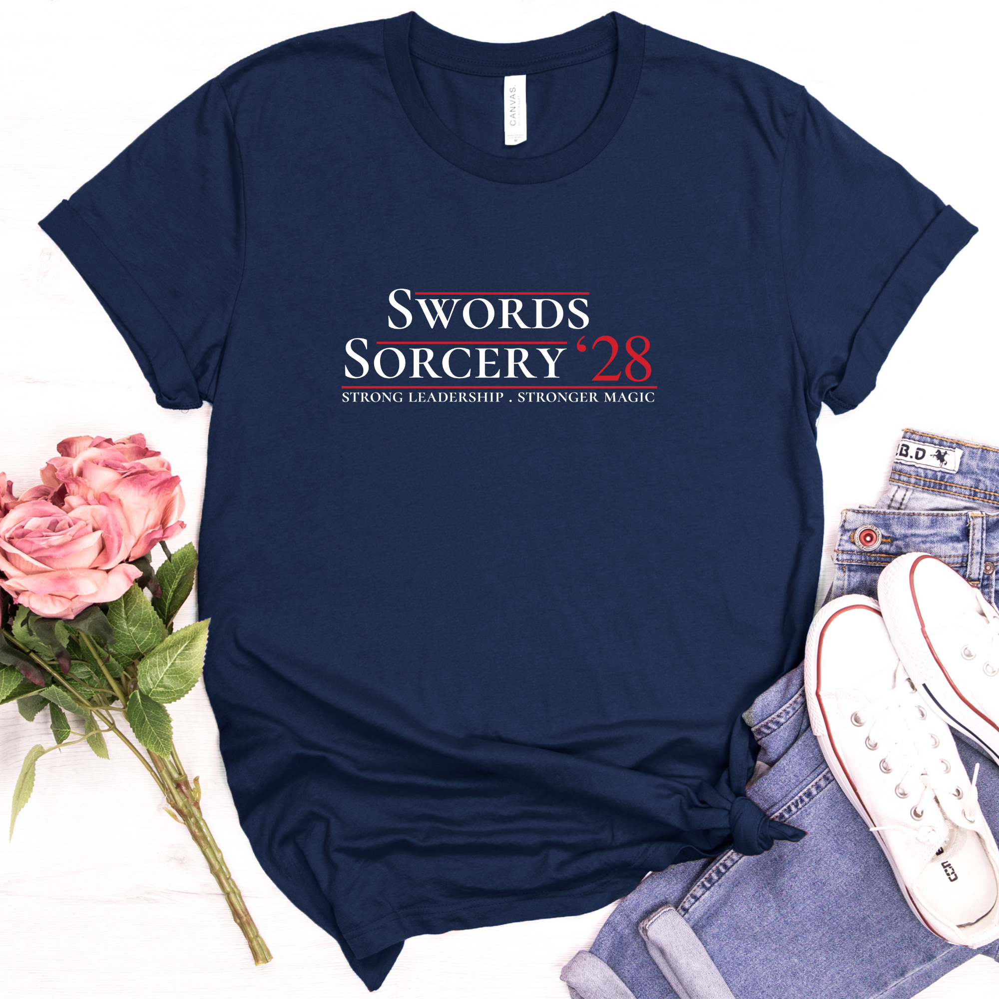 Styled flat lay of Swords Sorcery ’28 parody campaign T-shirt with jeans, white sneakers, sunglasses, and flowers on a light tabletop.