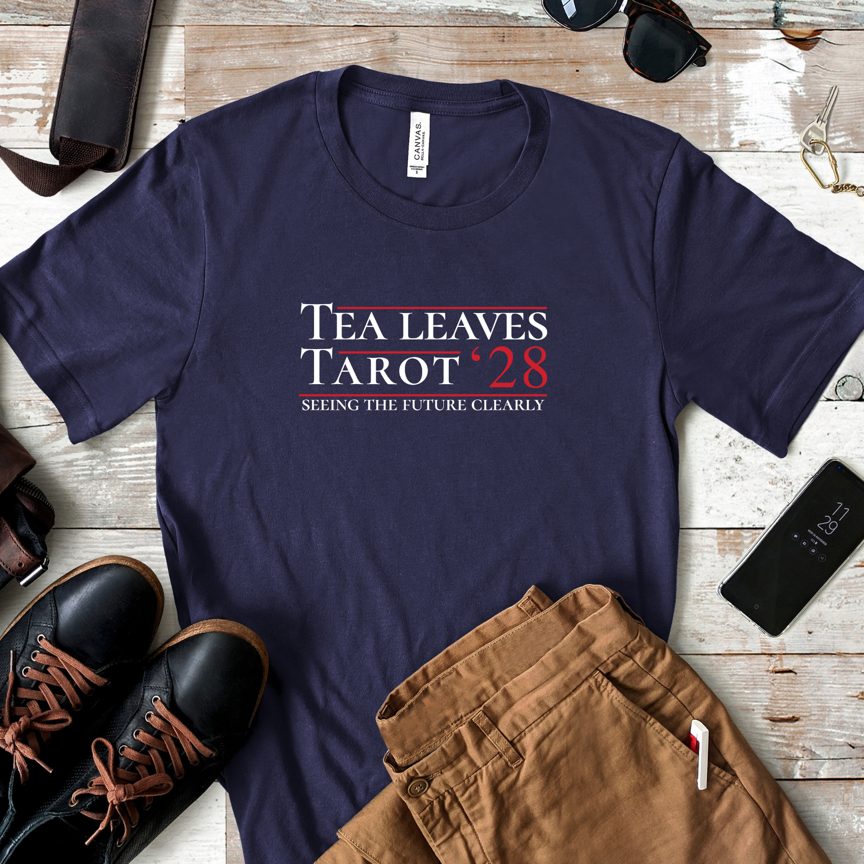 Flat lay of Tea Leaves Tarot ’28 parody campaign shirt on rustic wood background with boots, khaki pants, sunglasses, and accessories.