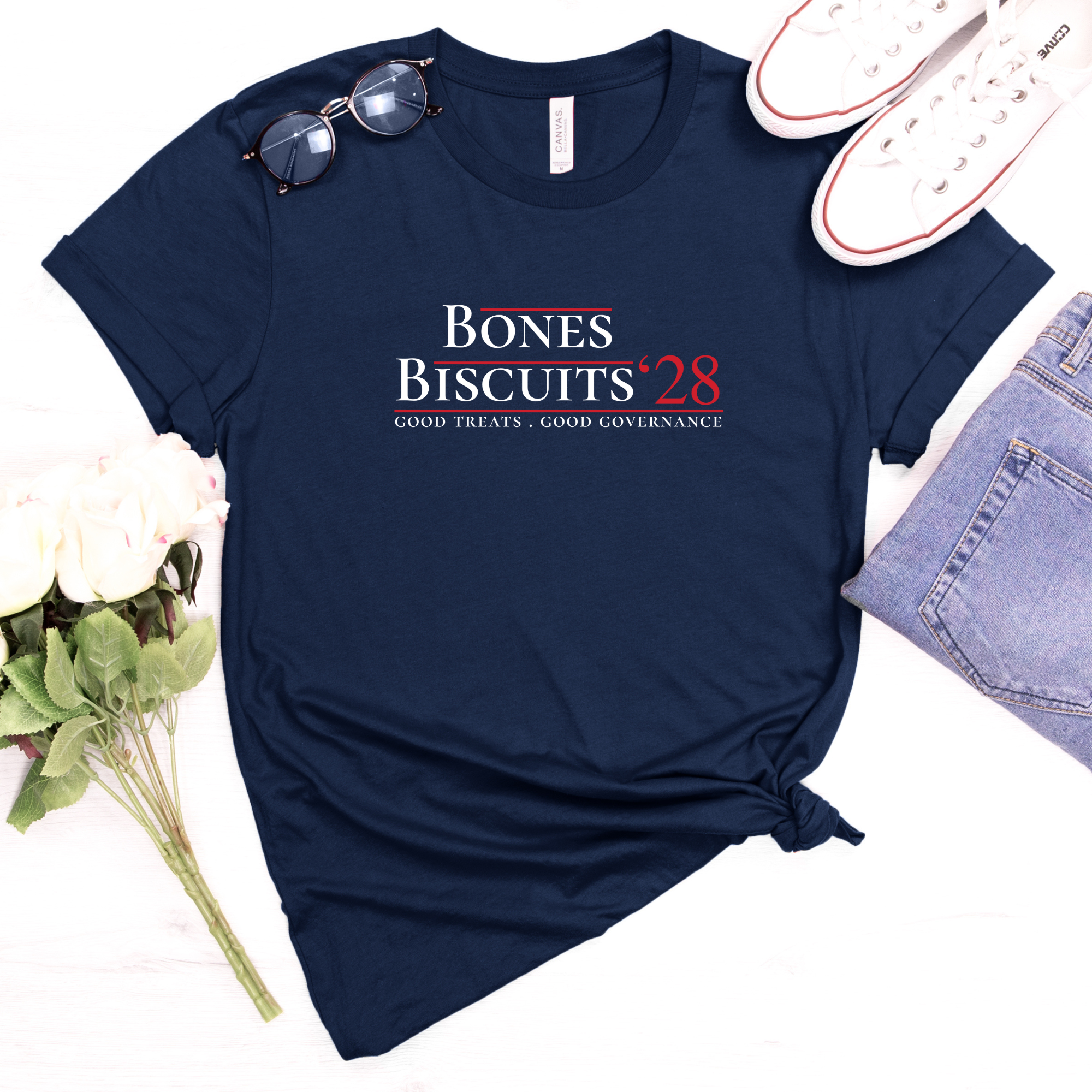 Navy Bones & Biscuits ’28 parody campaign shirt displayed with jeans, flowers, and casual accessories.