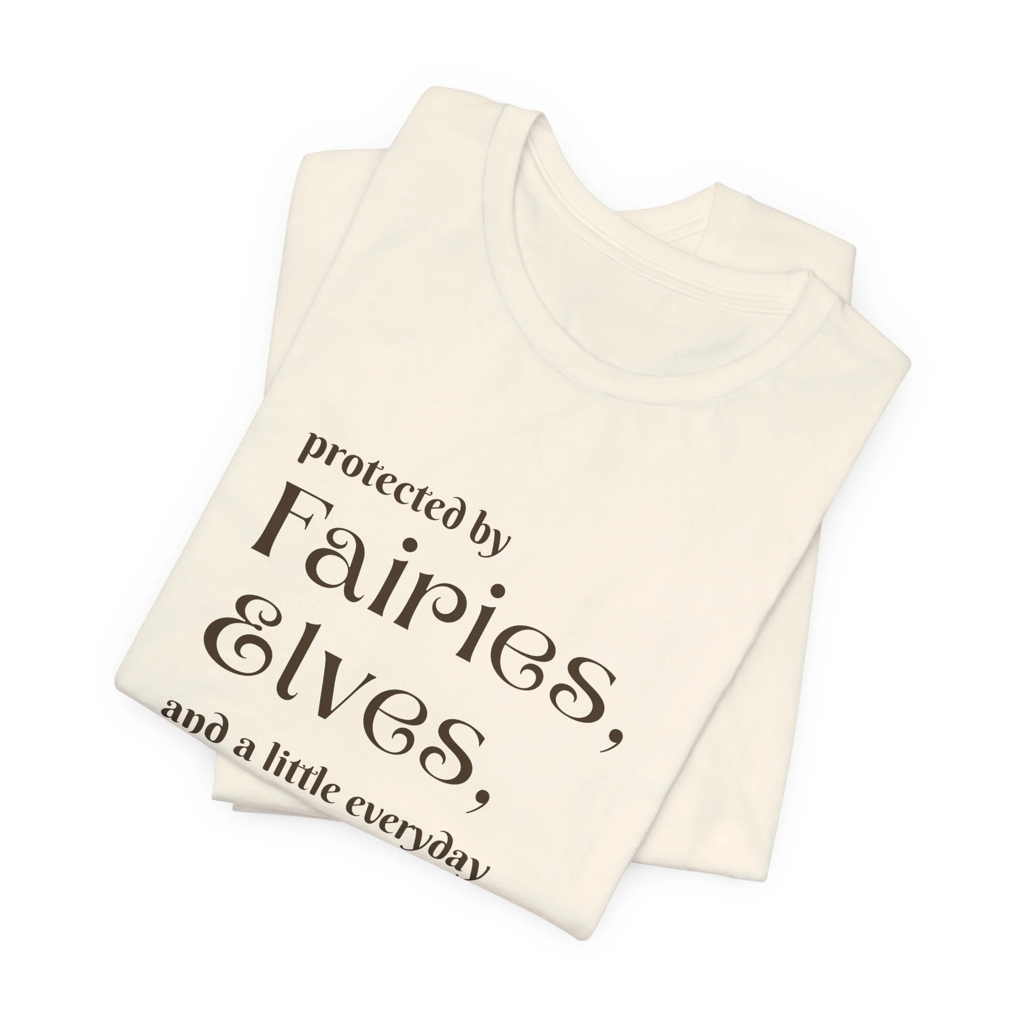 Folded natural-colored fantasy shirt with dark brown text reading protected by fairies elves and everyday magic