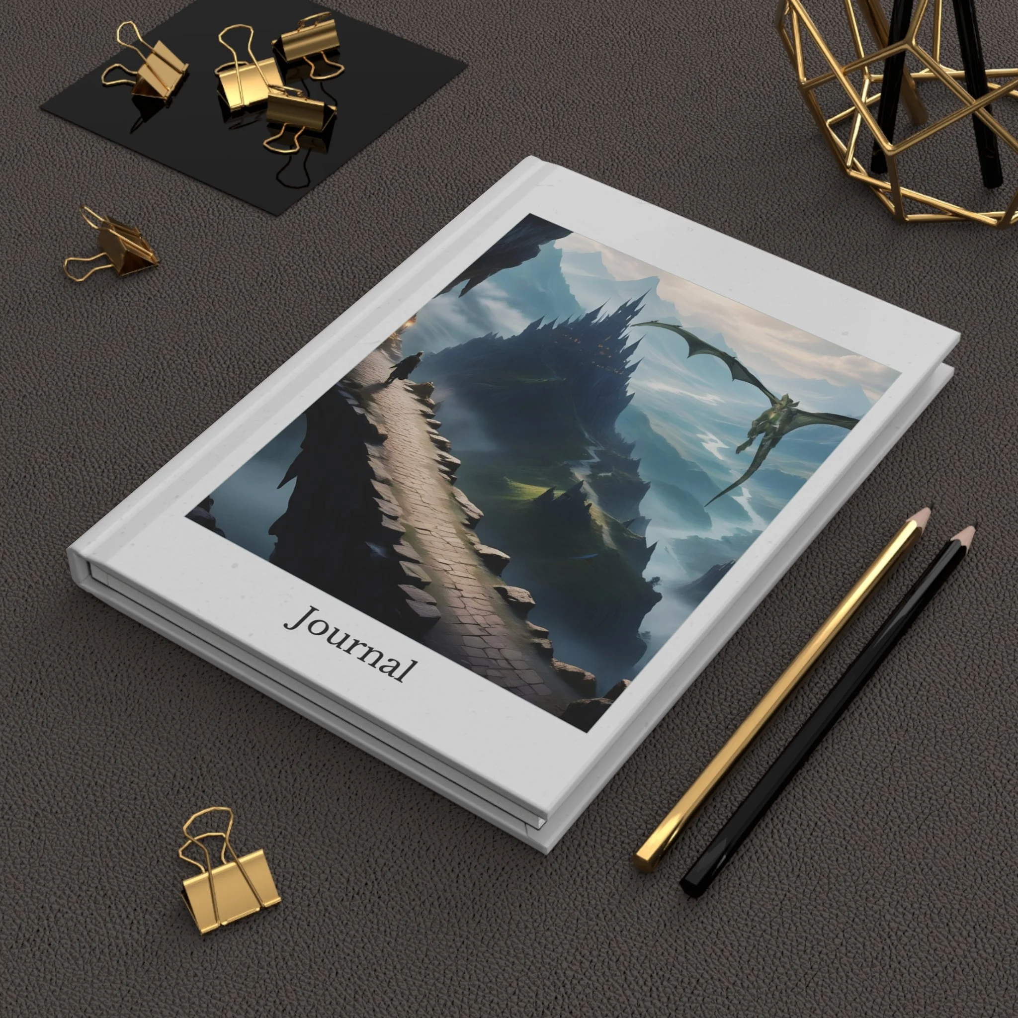 Angled desk view of “The Crossing” fantasy journal with stone bridge and castle artwork, shown with pencils and gold binder clips