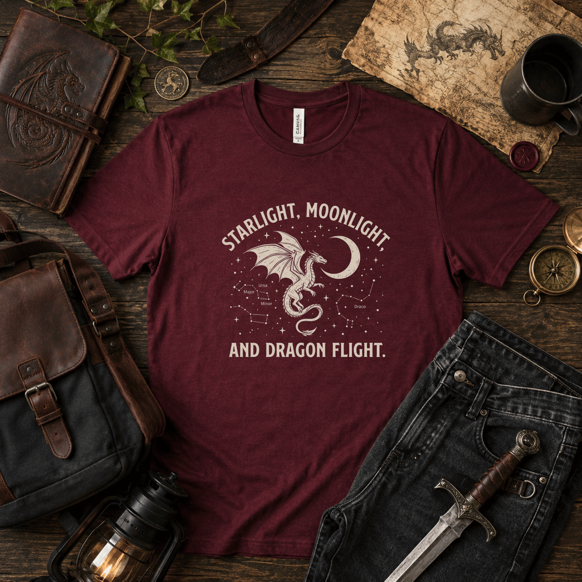 Lifestyle mockup of a Maroon Bella+Canvas fantasy dragon t-shirt styled with leather journals, a sword, maps, and other adventure-inspired props.
