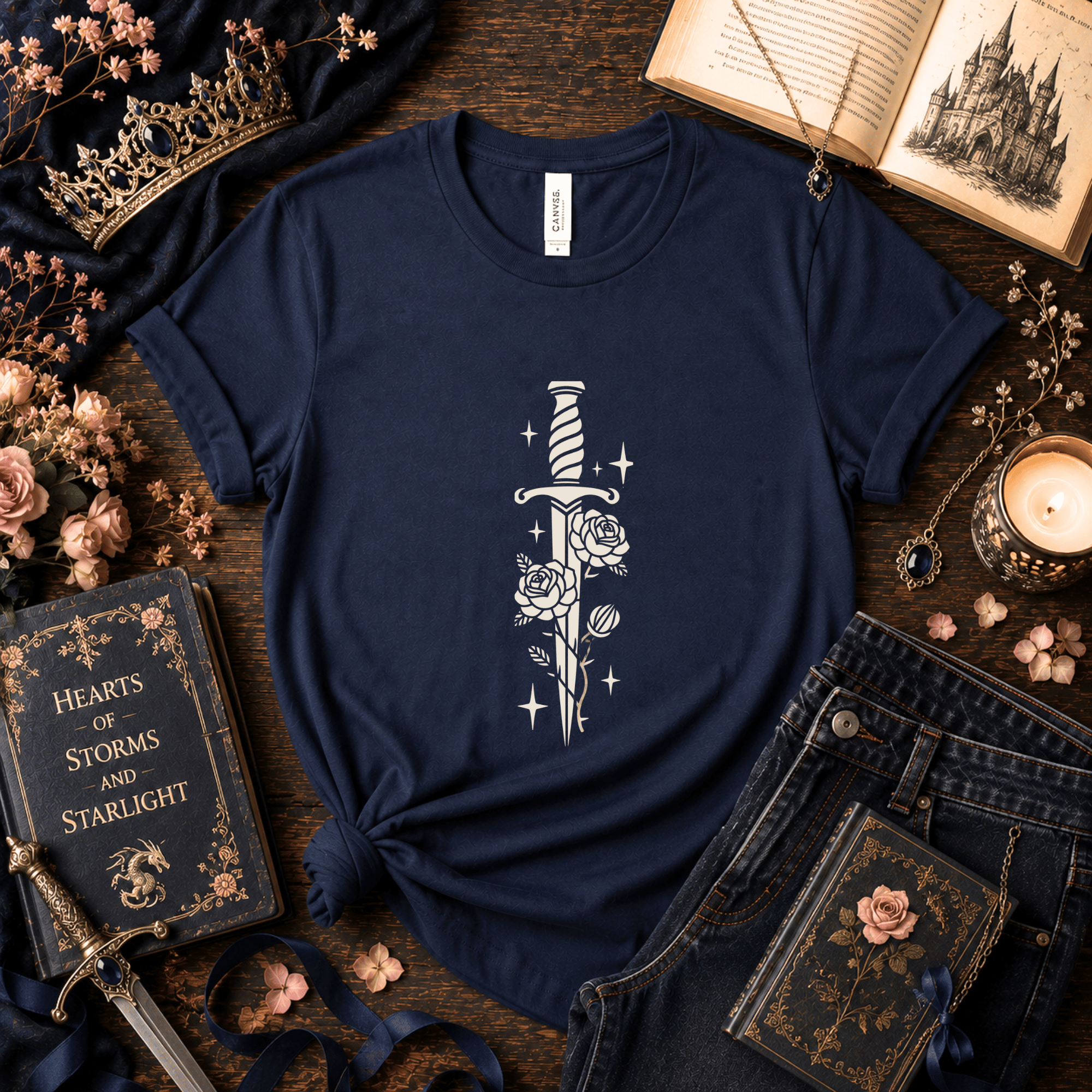 Lifestyle mockup of a Navy Bella+Canvas dagger and roses fantasy t-shirt styled with antique books, candles, florals, and dark academia-inspired accessories.