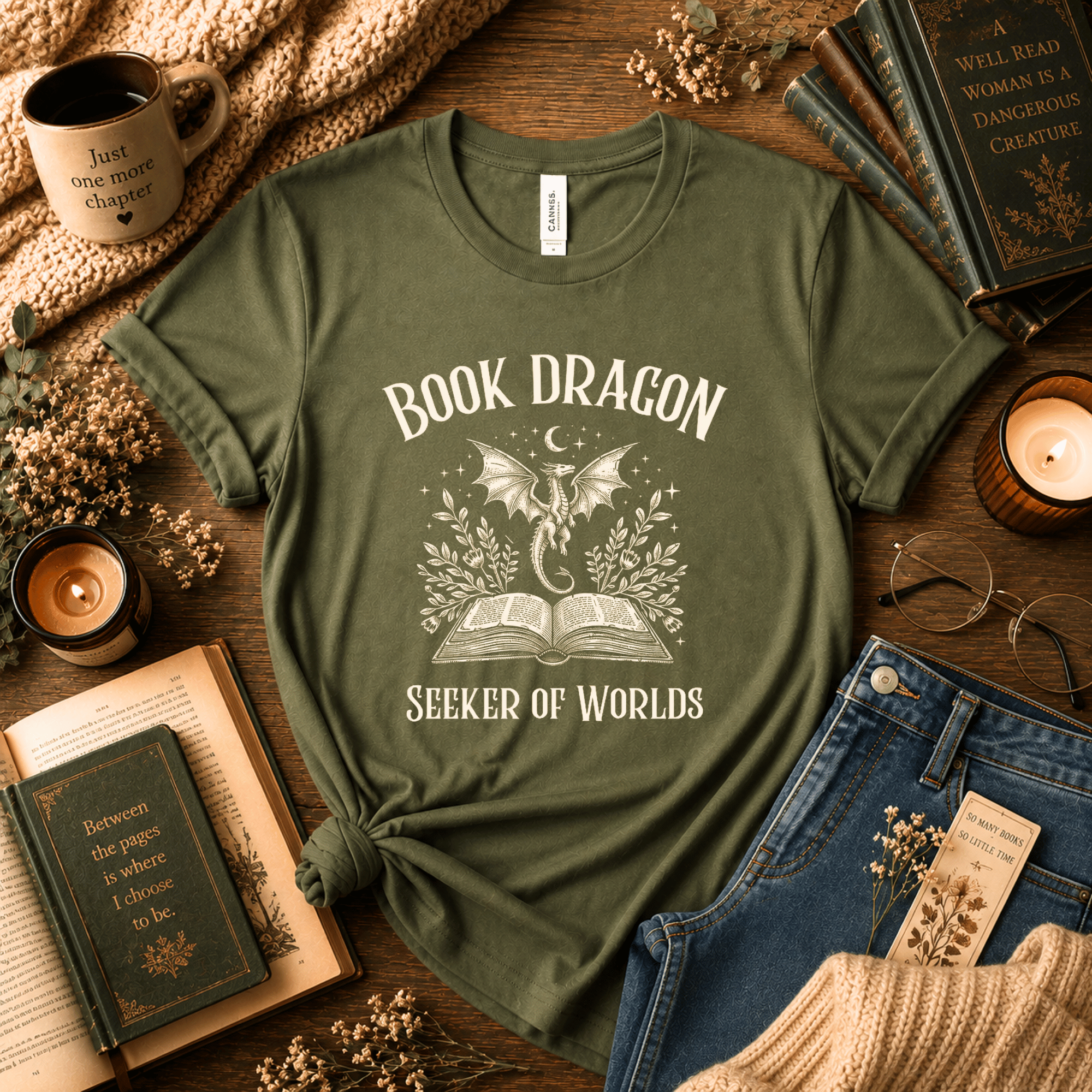 Lifestyle mockup of an Olive Bella+Canvas "Book Dragon" t-shirt styled with open books, candles, coffee, and cozy fantasy reading accessories.