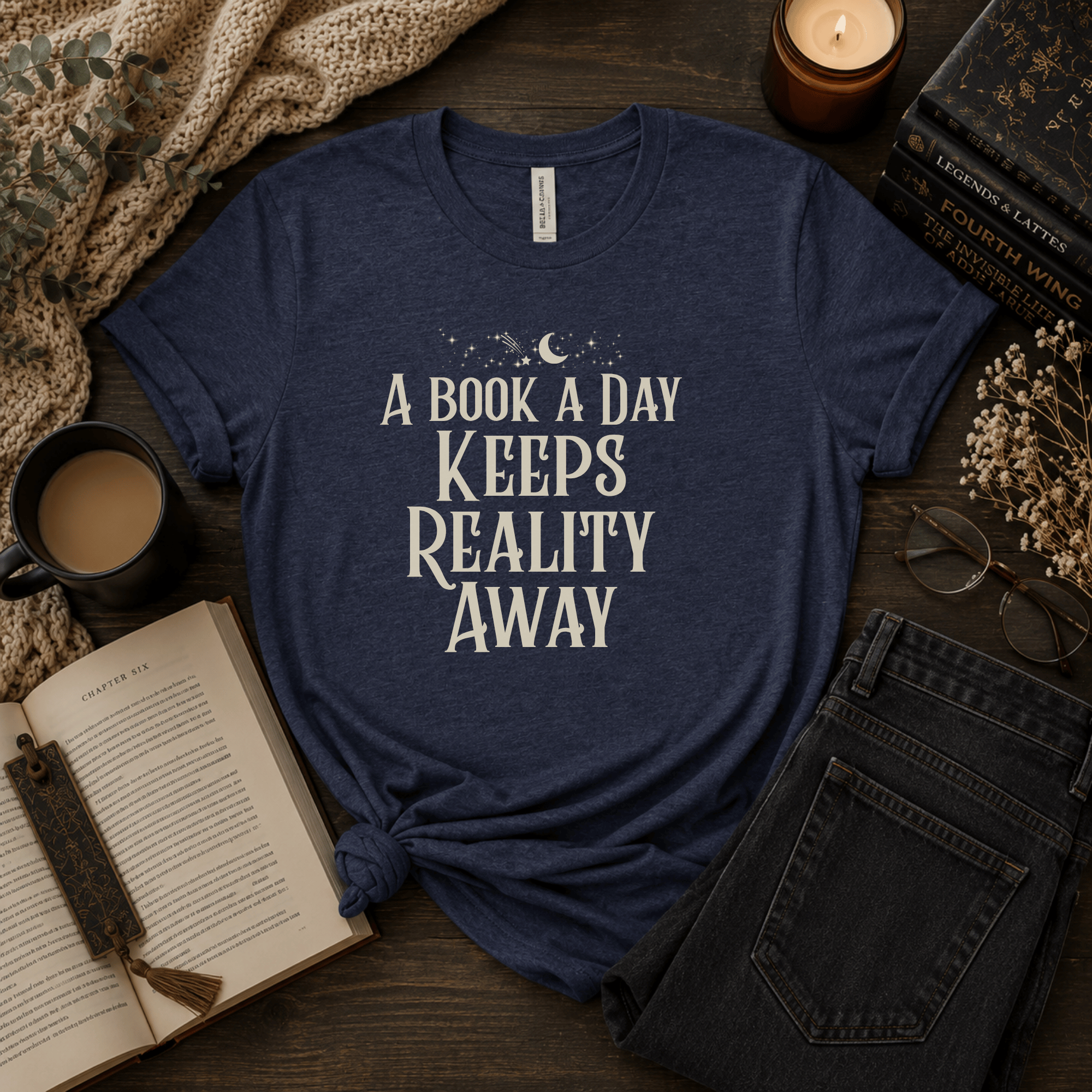 Lifestyle mockup of a Navy Bella+Canvas "A Book a Day Keeps Reality Away" t-shirt styled with an open book, coffee, candles, and cozy reading accessories.