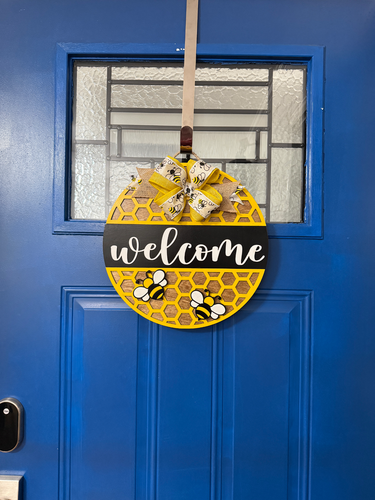 Front door display of honeycomb-pattern welcome door hanger with bees, black banner text, yellow bow, and greenery on a blue door