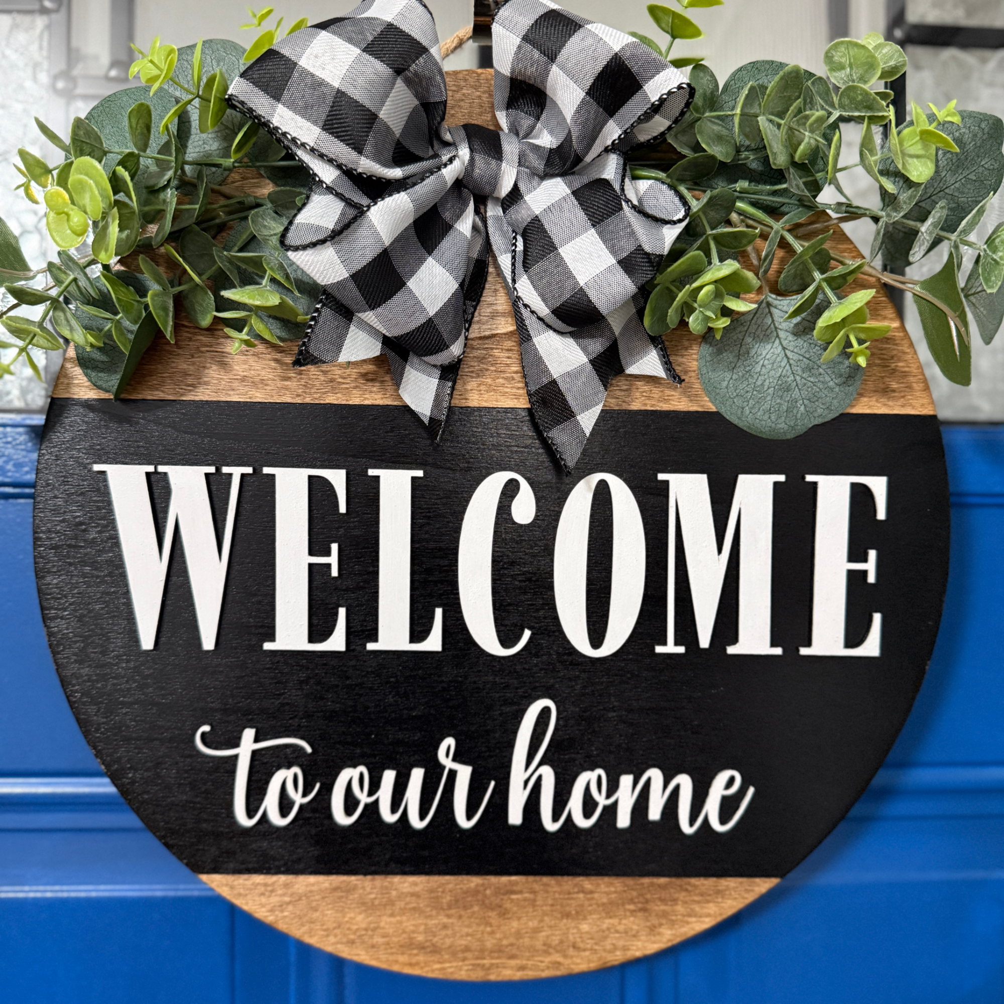 Black and natural wood welcome to our home door hanger with white lettering, greenery accents, and buffalo plaid bow on blue front door