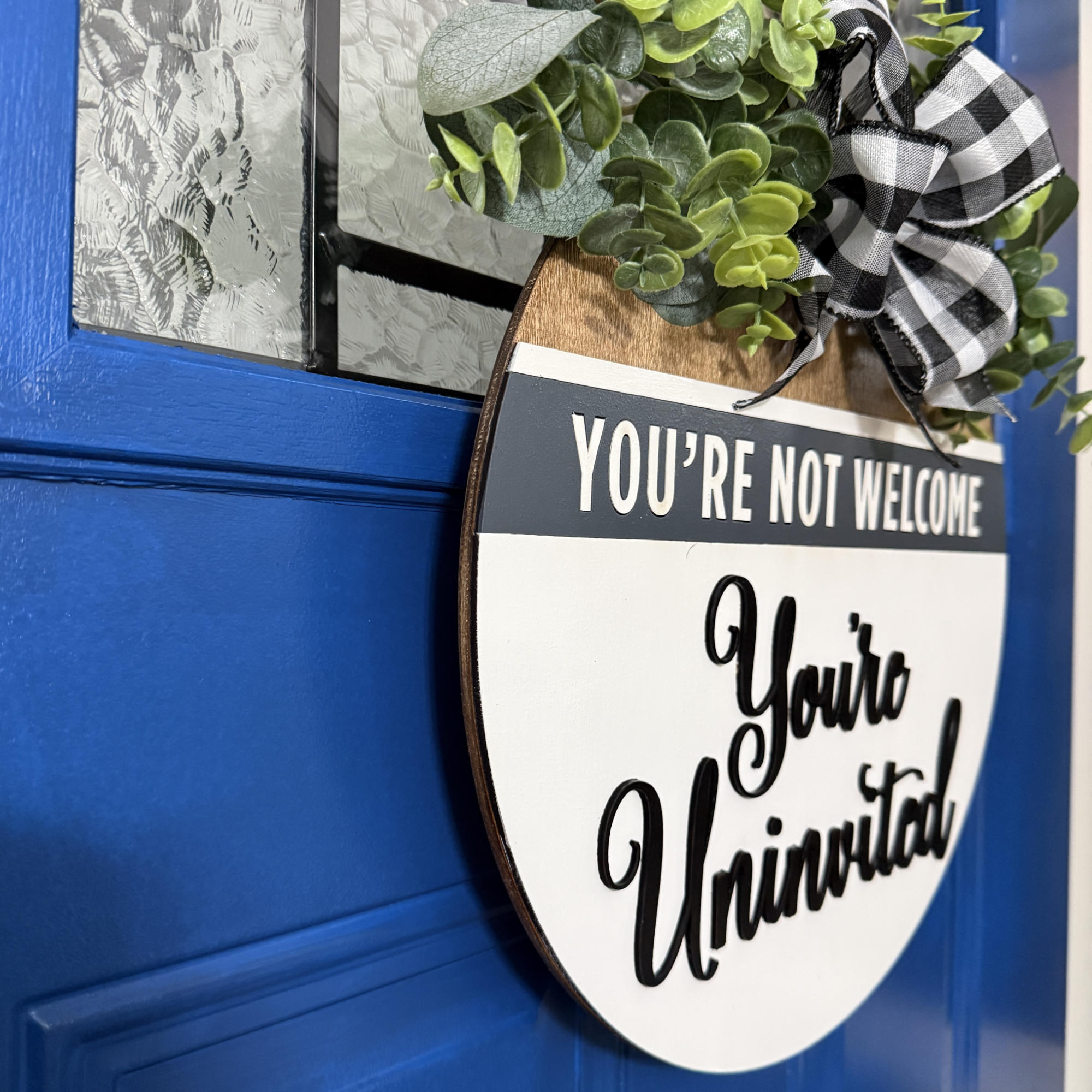Angled side view of white wooden “You’re Uninvited” door hanger showing layered dimensional lettering, gray top panel, and black and white buffalo plaid bow on a blue front door.