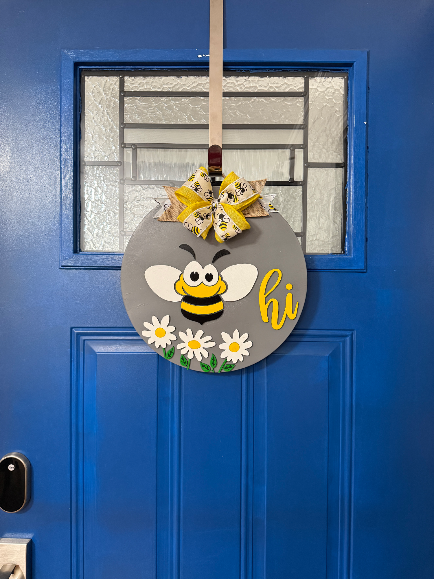 Front door display of gray round bee-themed door hanger with smiling bee, white daisies, yellow bow, and “hi” text on a blue door