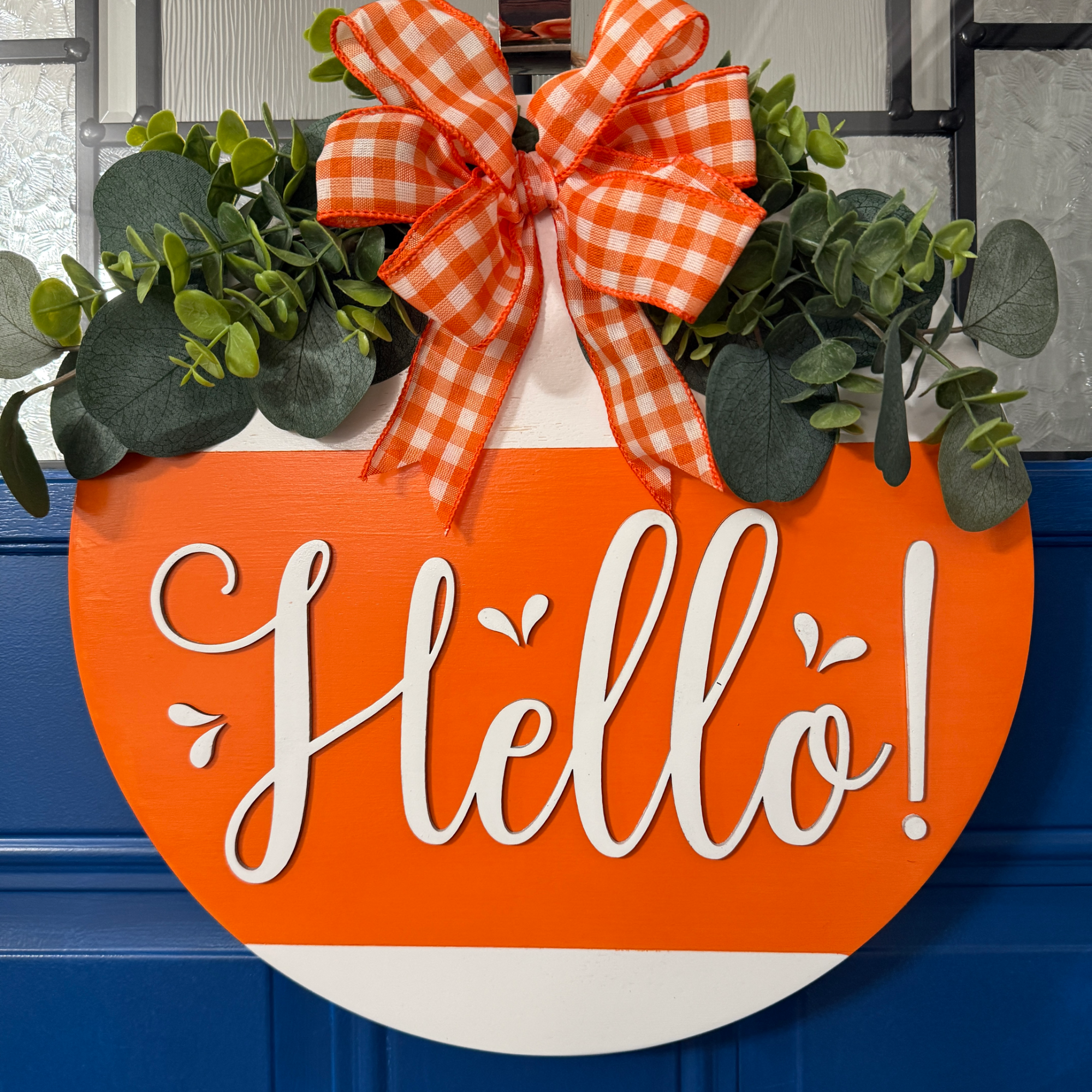 Orange and white seasonal Hello door hanger with greenery accents and orange gingham bow on blue front door