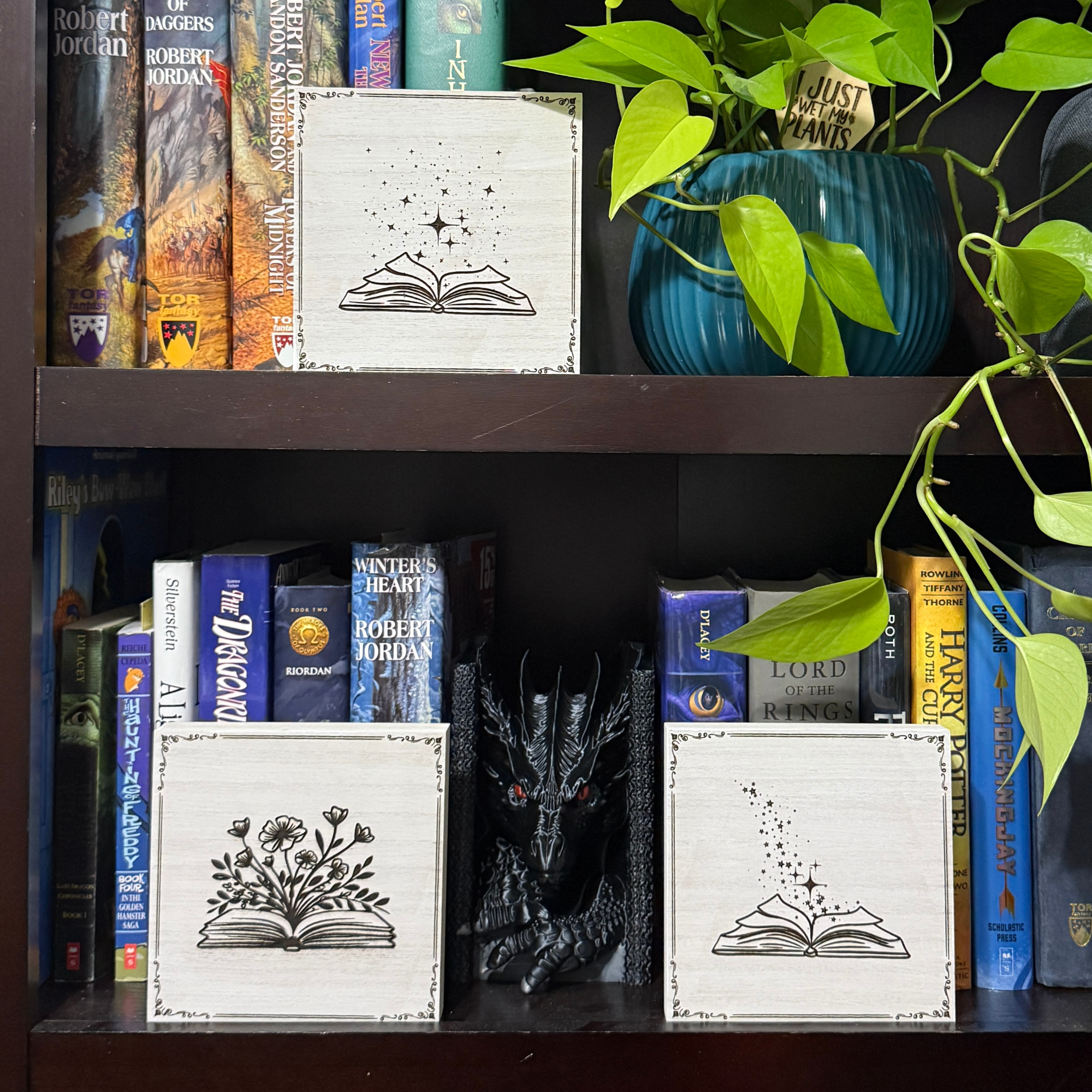 Fantasy book-themed shelf sitters displayed on a bookshelf, including magic and fairy book designs surrounded by novels and greenery