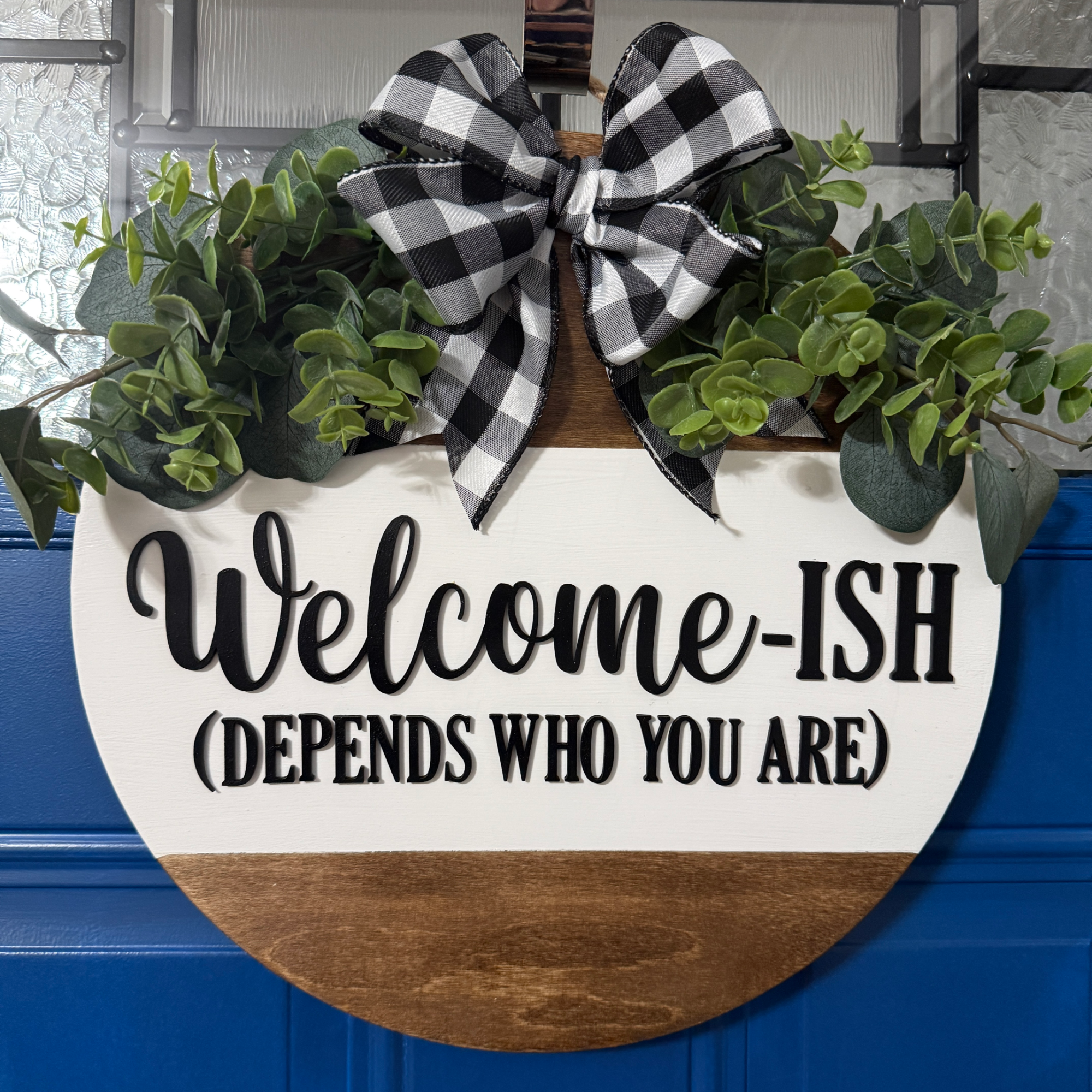 Front view of round wooden door hanger with white-and-natural wood finish, white text reading ‘Welcome-ISH (Depends Who You Are),’ buffalo check bow, and eucalyptus greenery.