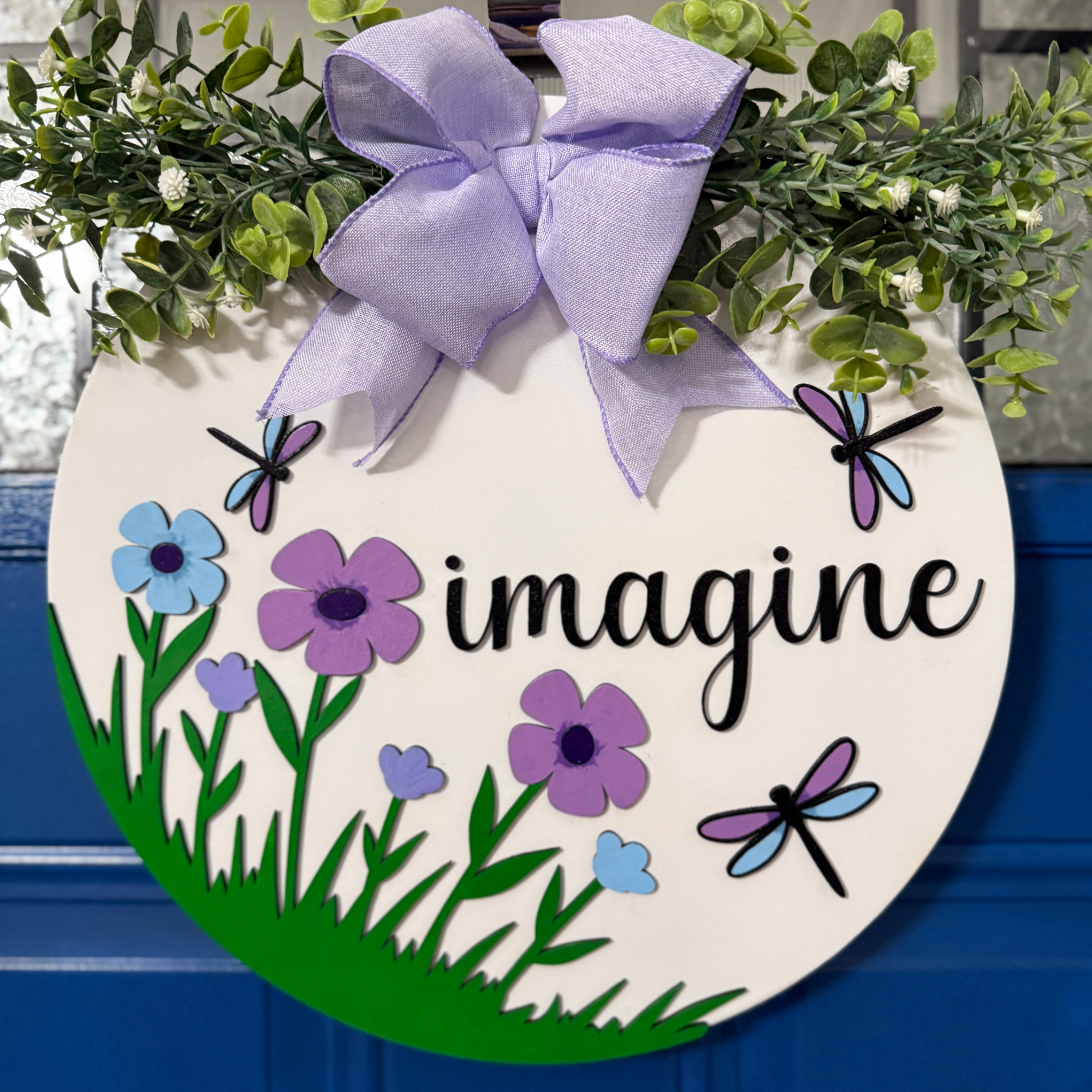 Front view of round floral door hanger with purple flowers, dragonflies, and “imagine” text, hanging on a blue door with lavender bow